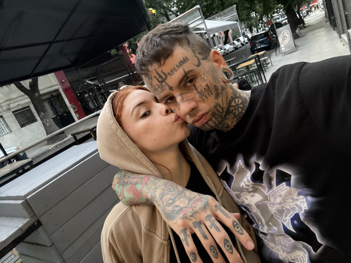 Two individuals are captured in a close-up selfie format on a city street in Buenos Aires, Argentina. The person on the left, identified as female, has reddish-brown hair and wears a light brown hooded sweatshirt with the hood up. Her left arm, which is extensively tattooed with colorful designs, rests on the shoulder of the second individual. She is seen with lips pursed, simulating a kiss towards the camera, and has a visible septum piercing. The person on the right, identified as male, has short dark hair and extensive facial tattoos, including a trident on his forehead, various symbols and text around his eyes, cheeks, and chin. A tattoo resembling a bandage is visible on his right cheek. He exhibits large ear gauges and neck tattoos. He wears a black t-shirt featuring a stylized white and blue design on the front. Both individuals are positioned closely together. In the background, an urban environment is visible, featuring an outdoor cafe seating area with dark tables and chairs under a black awning, a sidewalk lined with trees, and several parked vehicles along the street. Buildings with multiple windows are also present in the background.