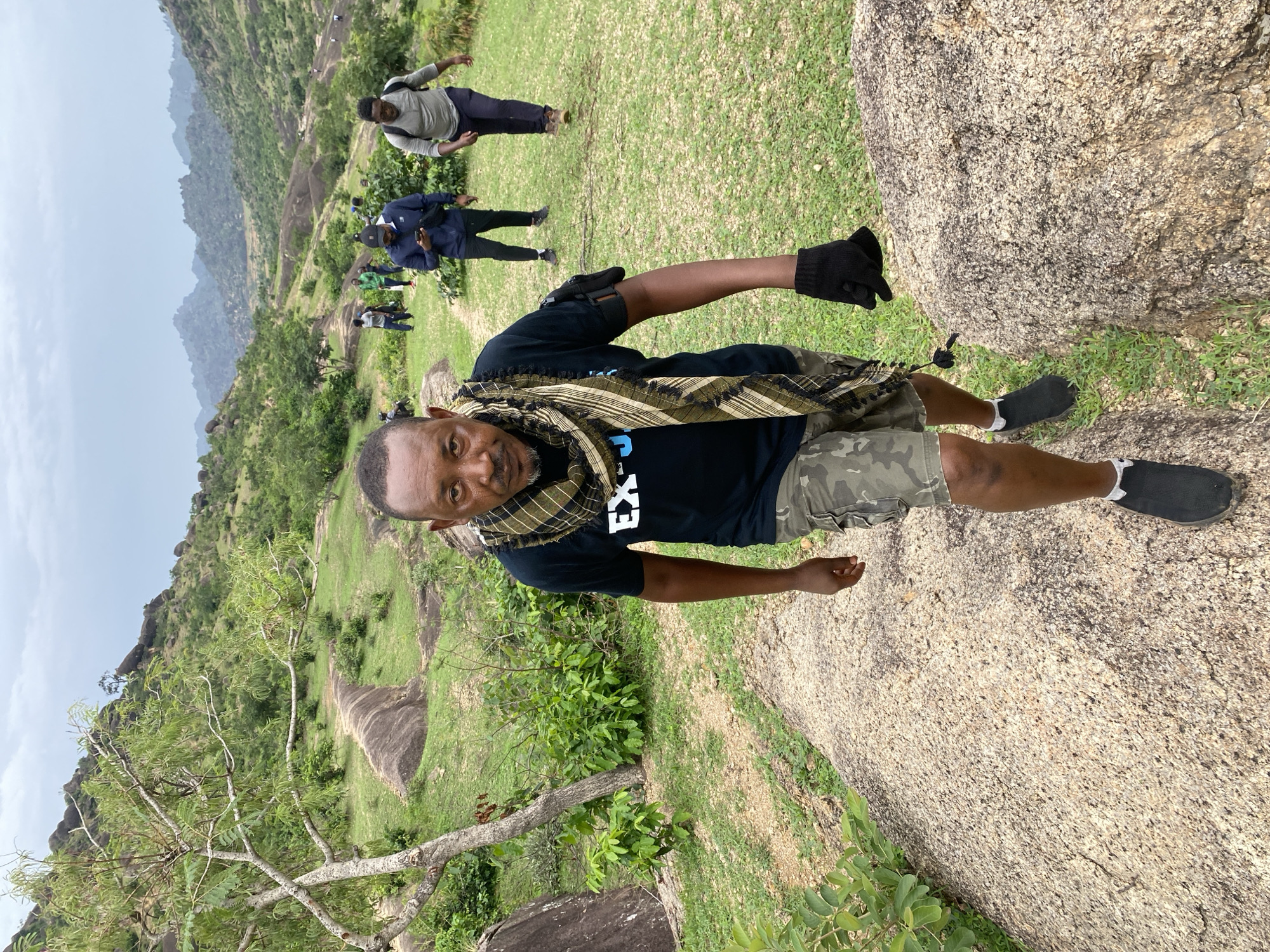The image shows a man in a black t-shirt, camouflage shorts, and black shoes standing on a large rock formation. He has a patterned scarf around his neck and a black glove on one hand. In the background, there is a landscape with other people, grass, trees, and more rock formations. The location appears to be a hilly or mountainous area. The image shows the location of Ganawuri, Nigeria.