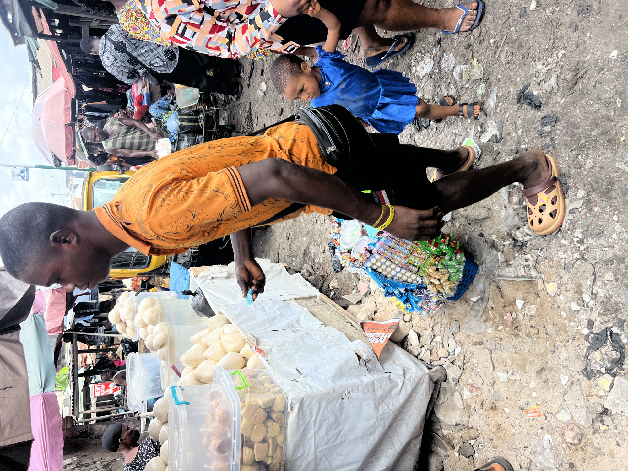 This image captures a bustling outdoor market scene in Nkpor, Nigeria, under an overcast sky during the daytime.
In the foreground, a man with dark skin, wearing an orange patterned short-sleeved shirt, black trousers, and light brown Crocs-style clogs, is bending over a market stall. He has a yellow wristband on his right arm, with partial text "ment" visible. He appears to be a vendor, either arranging or selling goods. The stall in front of him is covered with a white tarp and displays numerous clear plastic containers filled with rounded, light-colored food items, possibly fried snacks or pastries.
To the man's right, a small child in a bright blue dress and black sandals stands looking down. An adult, partially visible in a colorful patterned shirt, is holding the child's hand. Another person's feet, clad in blue flip-flops, are also seen.
The ground is unpaved, uneven, and consists of dirt, small rocks, and scattered litter, including plastic bags. Near the man's feet, on the ground, are small baskets and piles of individually packaged candies and snacks, some with text like "LAV" and "ments" visible. An orange and white discarded item nearby shows "ORE".
In the mid-ground and background, the market extends with various makeshift stalls and structures. A pink umbrella shades one stall that appears to be selling clothes. A person in a striped tank top is visible behind another stall. A yellow commercial vehicle, possibly a taxi or bus, is parked further back, and other blurred figures suggest a crowded, active environment.