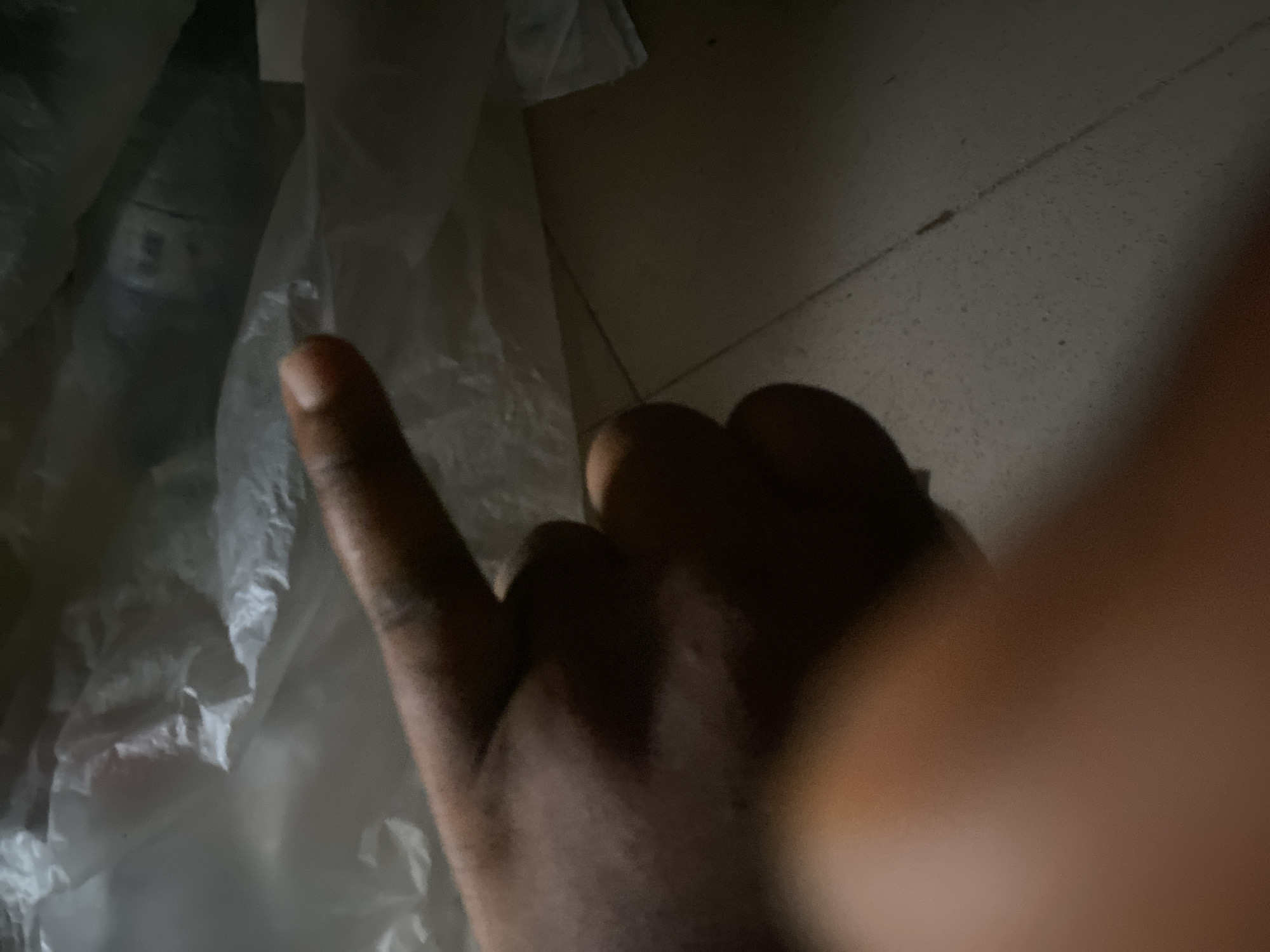 A close-up, low-angle shot focuses on a dark-skinned hand with its middle finger extended, pointing towards the upper right of the frame. The hand is in sharp focus, while the background is blurred and consists of white plastic material, possibly garbage bags. The floor beneath is a tiled surface, visible in the lower right quadrant of the image. The lighting is dim, creating deep shadows and highlighting the texture of the skin. The location, Jauro Yinu, Nigeria, is not visually depicted in the image itself; it is contextual information provided separately.