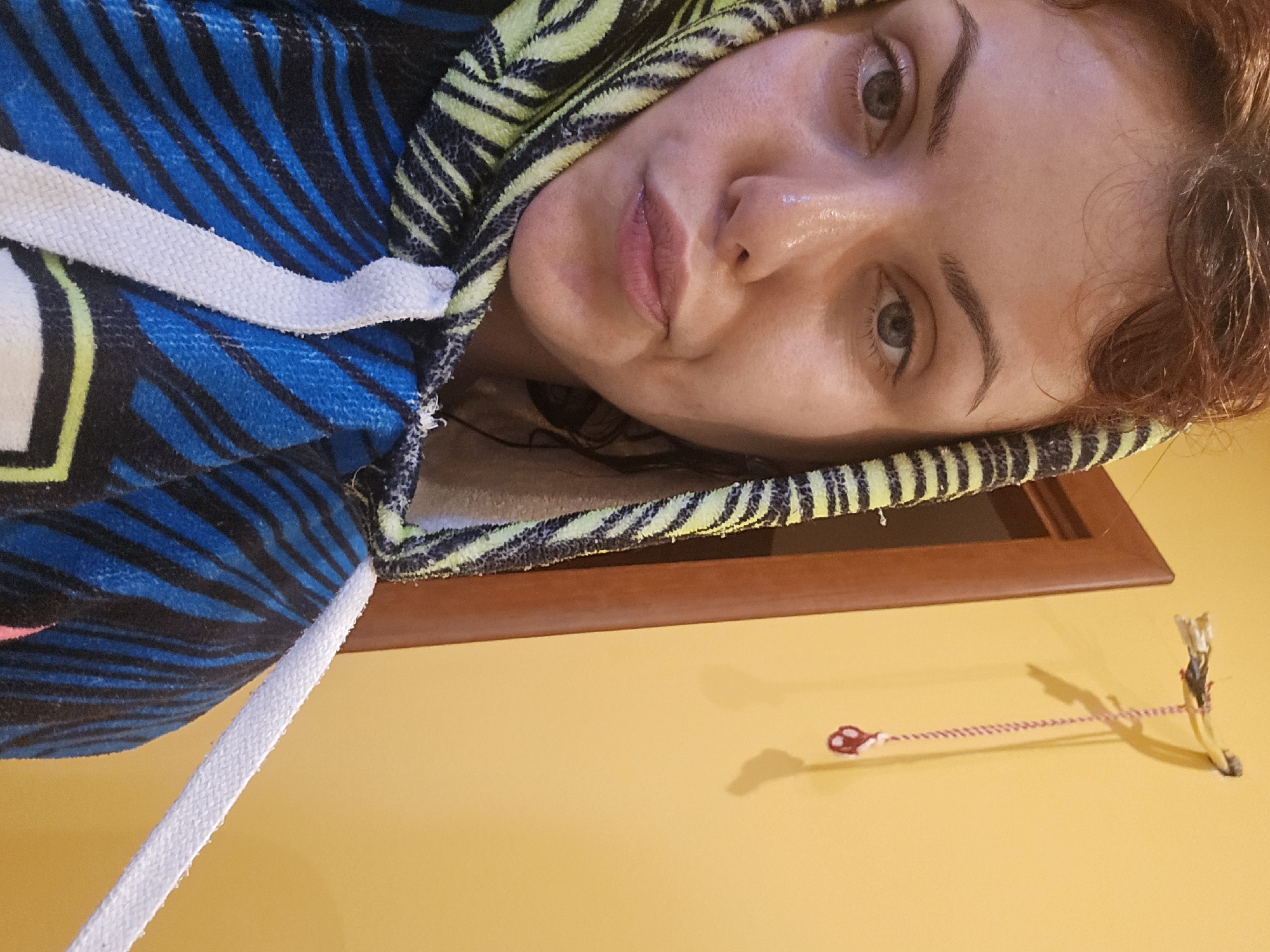 A woman, with reddish-brown curly hair, is captured in a close-up, front-facing selfie. She is wearing a hooded garment with a black, blue, and lime green zebra-like striped pattern. The hood is pulled up and draped around her face, and a white drawstring is visible on the right side of the garment. Her eyes are open and looking towards the camera, and her lips are slightly pursed. Her skin appears shiny, suggesting it might be sweaty or dewy.
The background of the image is an orange-yellow wall. To the left of the woman, part of a wooden doorframe is visible. On the wall, slightly above and to the right of the woman's head, a small, yellow object is attached, with a red and white braided string hanging down from it. The object appears to be some sort of decorative item or a part of an electrical fixture that has been repurposed. The lighting suggests an indoor setting. The location is identified as Unknown, Unknown.