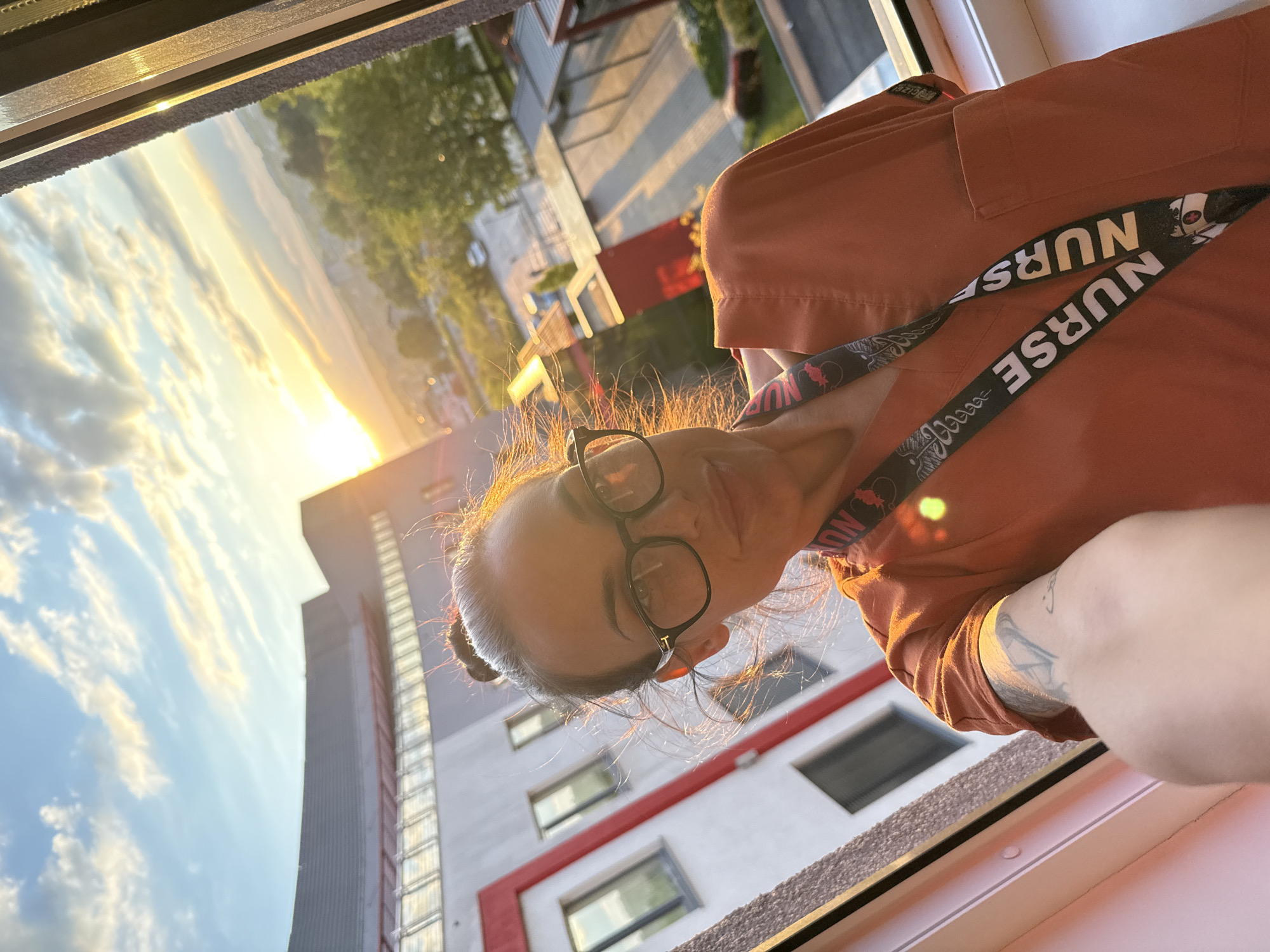 A woman with glasses and a "Nurse" lanyard is positioned in a window, overlooking a cityscape with a sunset in Pleven, Bulgaria. A multi-story building is visible outside the window. The woman is wearing an orange shirt. A tattoo is visible on her left arm.