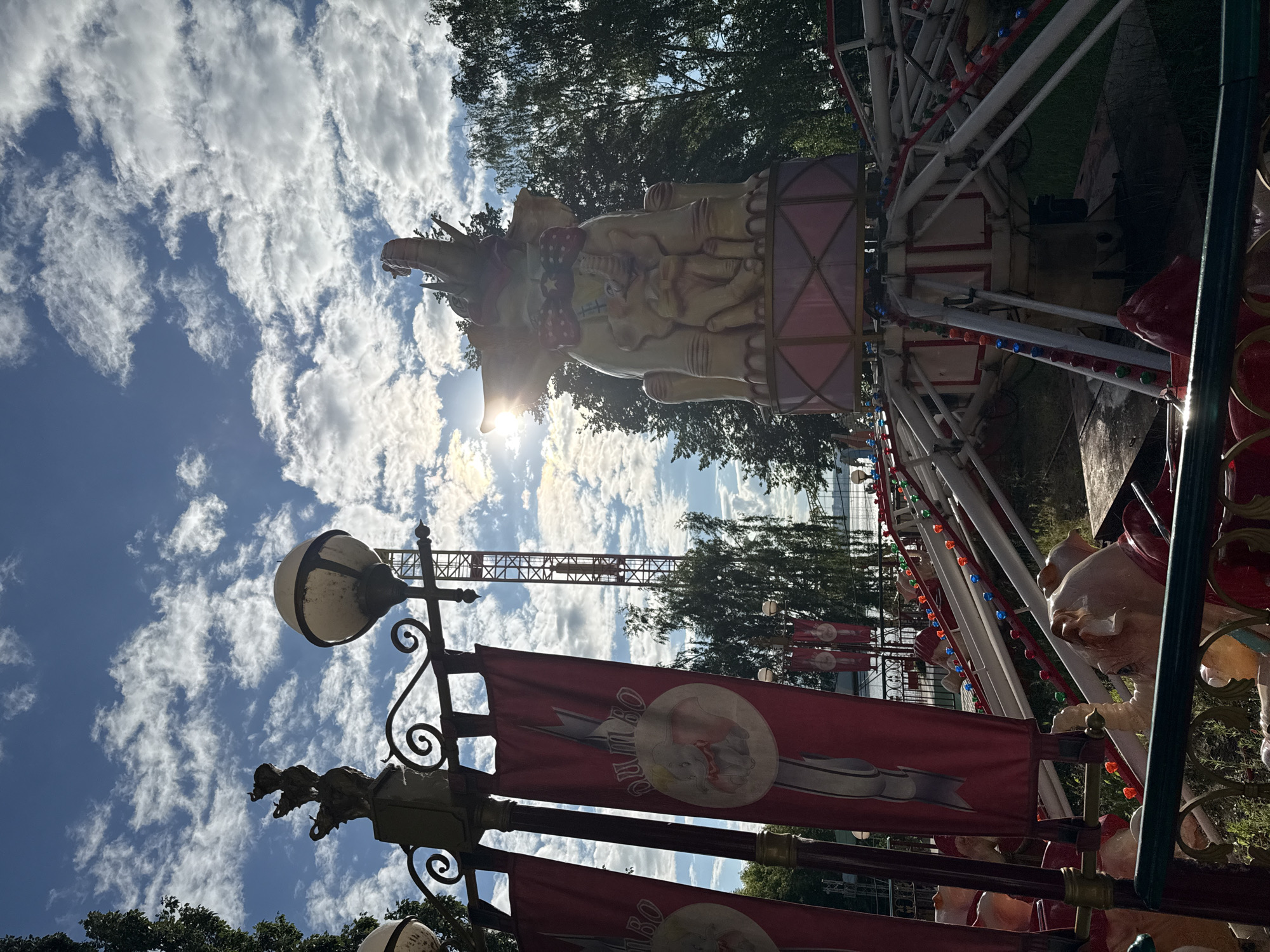 The image depicts an outdoor amusement park attraction, likely a carousel or a similar ride, with a Dumbo the flying elephant theme. In the foreground, a decorative fence is visible, with large, pale pink pig-like figures attached to the ride structure behind it. To the left, two red banners with a Dumbo illustration and Russian text are hanging from a pole.
Dominating the center of the image is a large, yellow, elephant-shaped sculpture, also themed around Dumbo, with a baby elephant on its back, adorned with a red bow tie. This sculpture sits atop the amusement park ride's machinery, which features a pink and white base and numerous mechanical arms and supports, some decorated with colorful lights.
In the background, a tall, red metal tower structure stands against a bright blue sky filled with white, fluffy clouds. The sun is visible through the clouds, casting a lens flare. Trees with green foliage line the background, suggesting a park or wooded area. In the far distance, a body of water and possibly distant buildings are faintly visible. The overall setting appears to be a sunny day in an amusement park. The image does not contain enough information to determine the specific location within the city of Staryi Senteg, Russia.