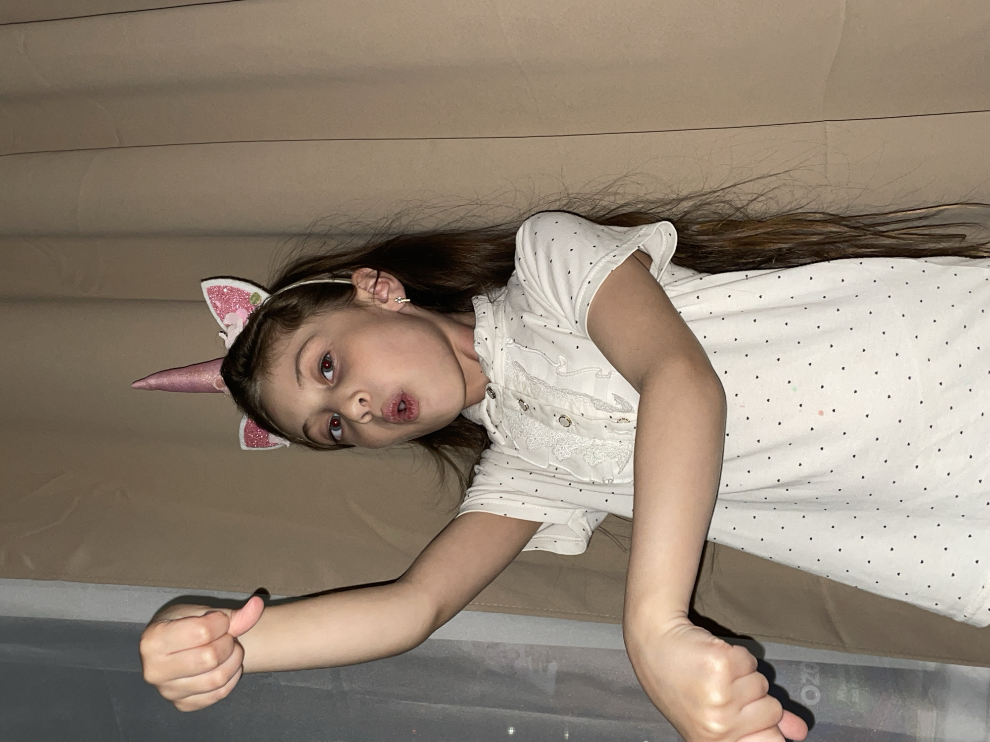 A young girl with brown hair, wearing a white polka-dot dress and a pink unicorn headband, is lying on a surface with her head tilted. Her left arm is extended and making a thumbs-up gesture. The background consists of a brown curtain. There is no indication of the city of Izhevsk, Russia in this image.