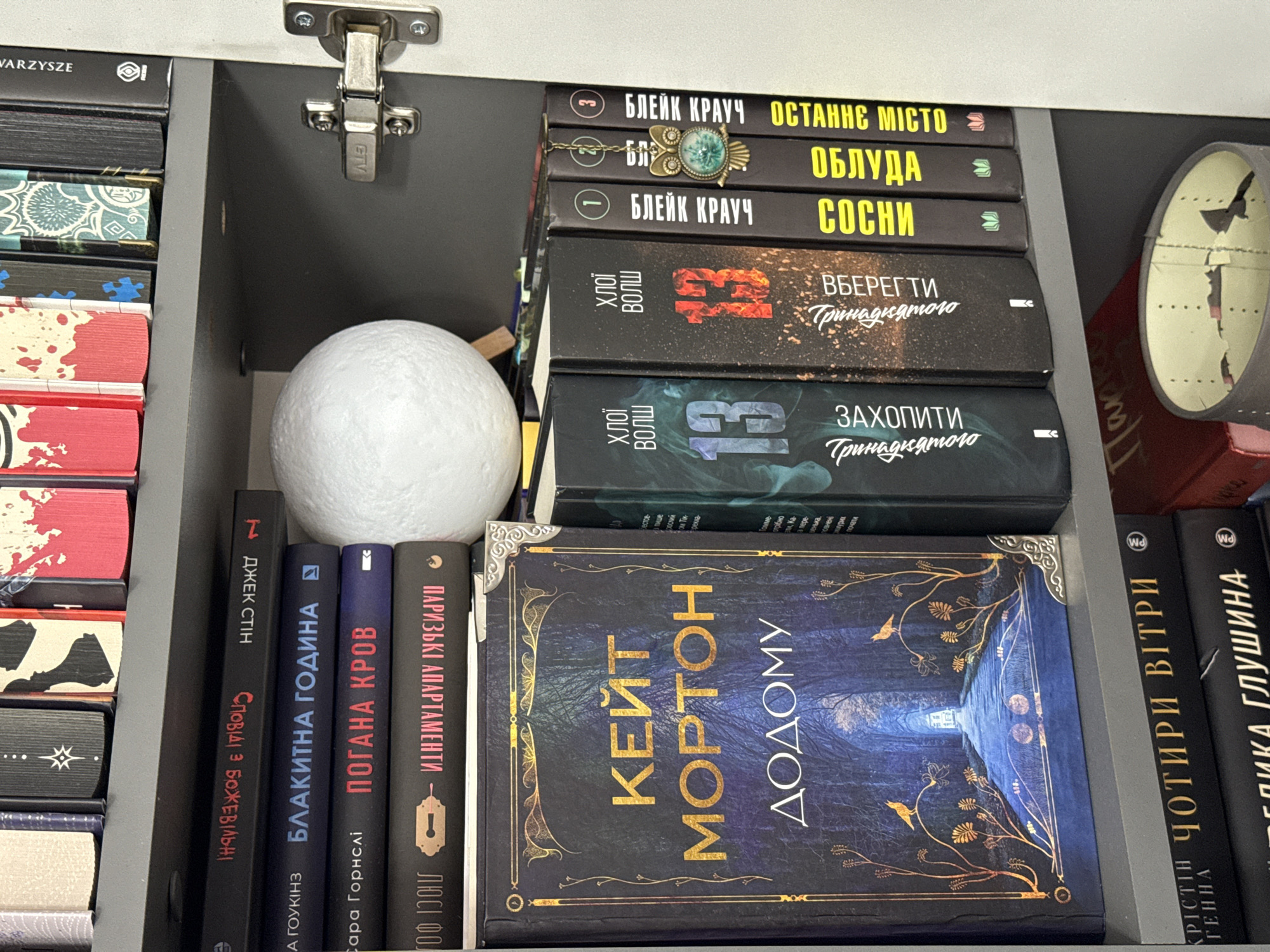 The image displays a section of a bookshelf filled with books and a decorative item. The bookshelf is dark gray, and the books have various colored spines. On the top shelf, several books are visible, some with brightly colored covers. One book has a black spine with white text.
The middle shelf contains a prominent book with a dark blue cover featuring a pathway leading into a forest. The title "КЕЙТ МОРТОН ДОДОМУ" is written in gold lettering on the cover. To the right of this book are several other books with black spines and yellow or white text. One of these books has a small owl-shaped charm attached to it. A white, spherical object, possibly a decorative moon, is also present on this shelf.
The bottom shelf has more books, with visible titles in white and yellow text. A portion of a red book and a grey cylindrical object with a broken lid are also visible on this shelf.
The scene appears to be an indoor setting, likely a room with a bookshelf. There are no people or actions depicted. The lighting suggests an indoor environment, and the overall impression is one of an organized collection of books.
There is no information in the image that indicates the location within the city of Łódź, Poland.