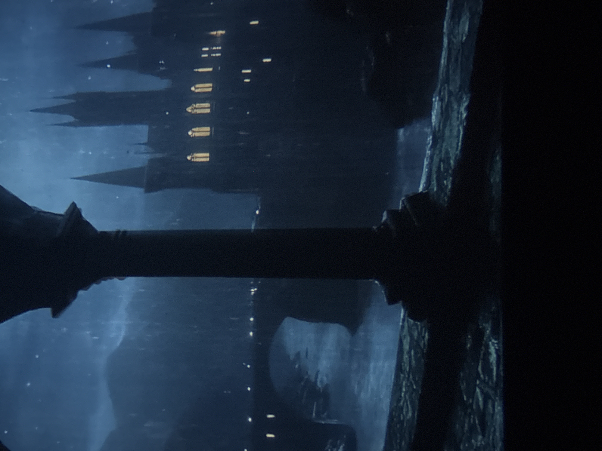 The image depicts a dark, nighttime scene featuring an architectural structure with castle-like elements. The predominant color is dark blue, and there are light yellow points scattered across the scene, likely representing illuminated windows. In the foreground, there is a dark, cylindrical object or structure, possibly a balustrade or a part of a building. The right side of the image is shrouded in darkness, showing the rough texture of a stone wall or cliff face. The architectural structure resembles a castle with several turrets and towers, typical of medieval or fantasy-inspired designs. The reflection in water is visible. The image does not appear to be a view of Varna, Bulgaria.