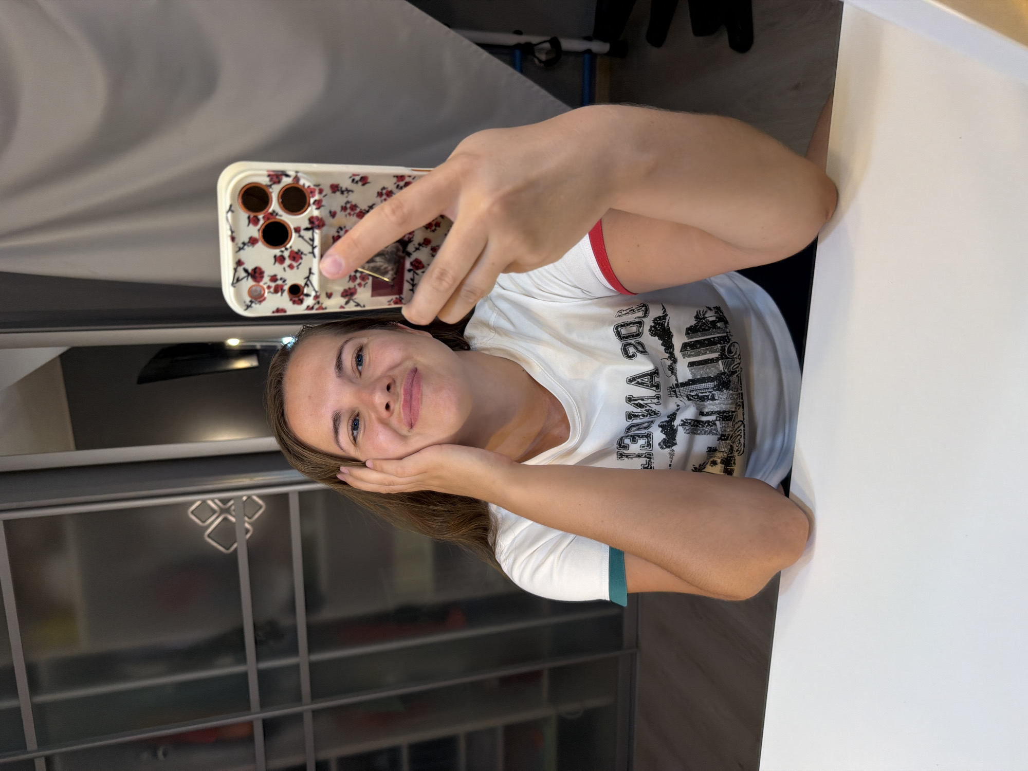 A young Caucasian woman with light brown hair is taking a selfie in a well-lit room. She is smiling and making a peace sign with her right hand. She is wearing a white t-shirt with a graphic design of a city skyline and palm trees. The t-shirt has green trim on the left sleeve and red trim on the right sleeve. Her left hand is resting on her cheek. She is seated at a white table, with her elbows resting on the table. In the background, there is a closet with mirrored doors and a light-colored curtain. The scene does not provide any specific indicators or landmarks to place it in Sochi, Russia.