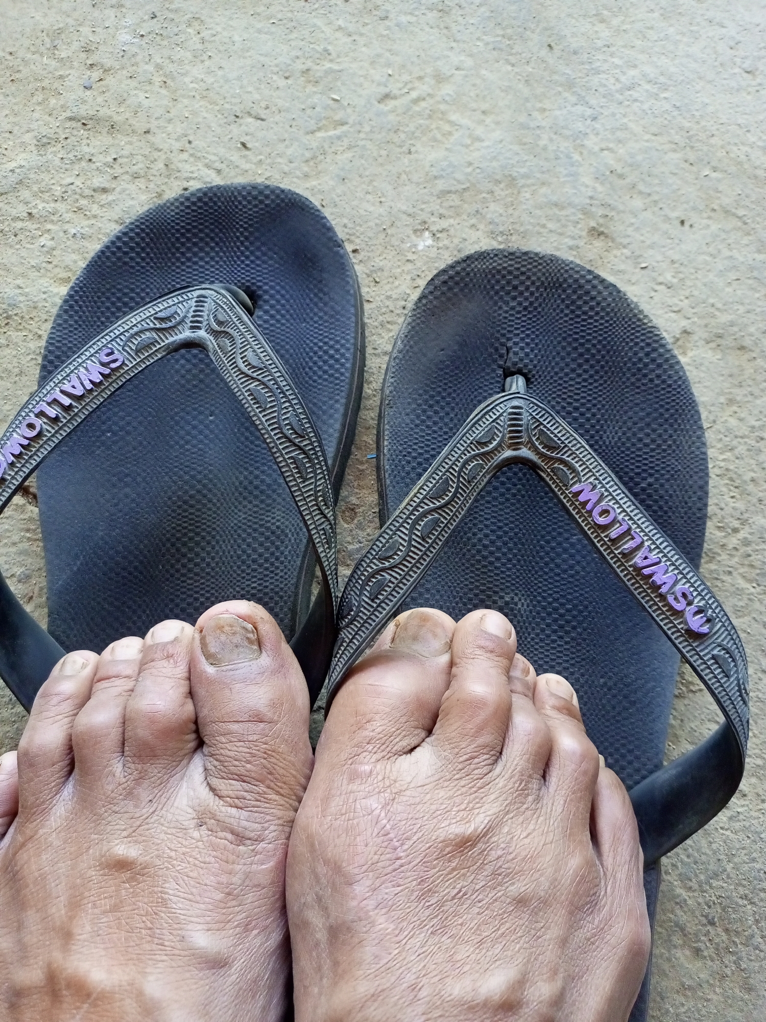 The image shows a close-up view of a person's feet wearing black flip-flops. The flip-flops have textured soles and straps with the word "SWALLOW" in purple lettering. The feet appear to be bare and are resting on a sandy or gritty surface, possibly a path or ground in an outdoor setting. The nails of the toes are visible, and the skin on the feet shows texture and some discoloration. The background is a neutral, textured surface that appears to be ground or pavement. The scene is consistent with an outdoor location in a warm climate, such as Jatipurno, Indonesia.
