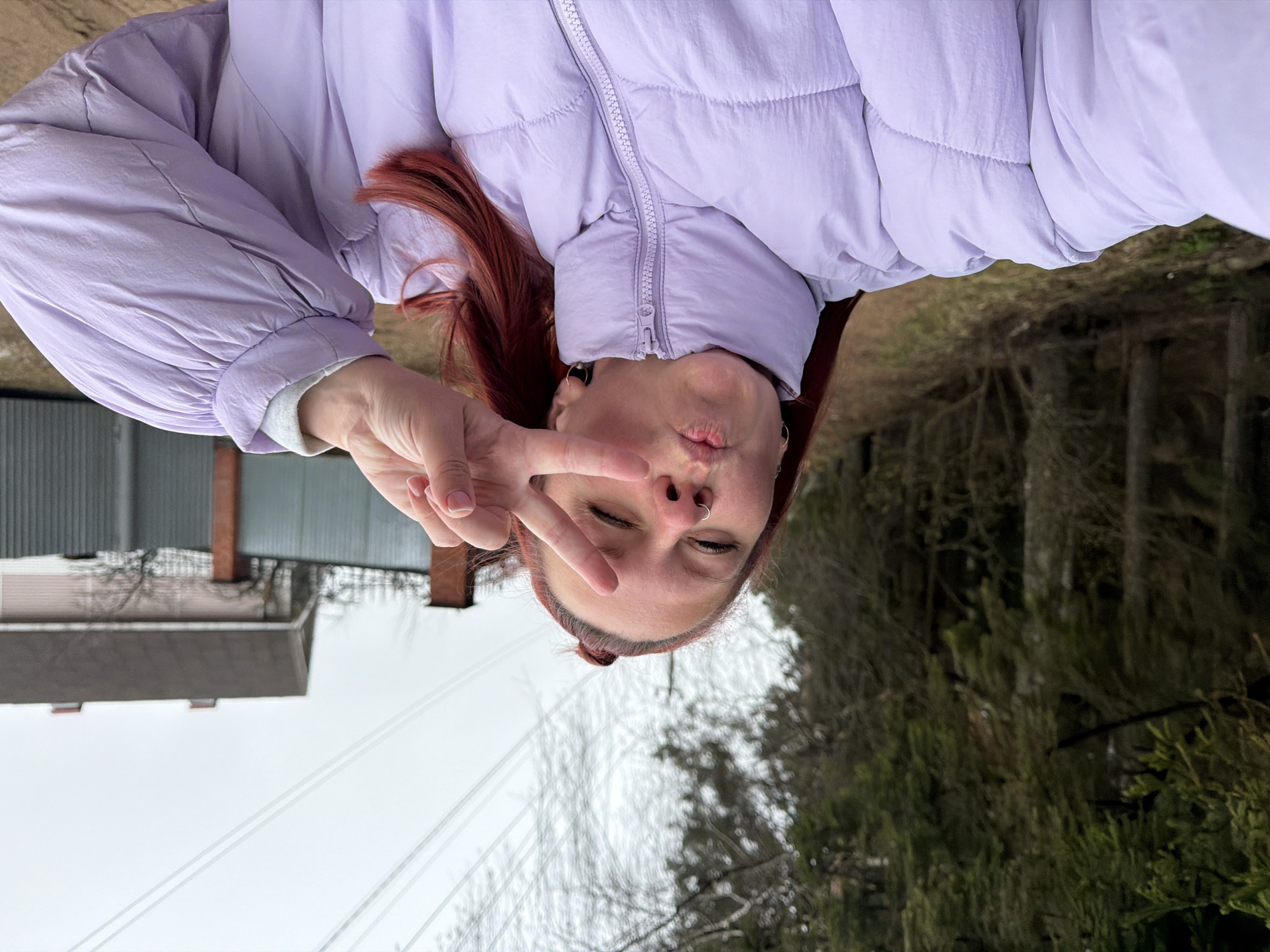 This rotated selfie-style image features a young adult female, appearing upside down as if the photo was taken with the camera inverted or subsequently rotated 90 degrees clockwise. She has reddish-brown hair and is wearing a light purple (lavender) puffer jacket, which is zipped up. A silver hoop nose ring is visible in her left nostril, and she has small earrings. Her right hand is raised, forming a peace sign, with her index and middle fingers extended, partially covering her closed or squinting eyes. Her lips are playfully pursed in a 'kissing' or 'duck face' expression.
The scene is outdoors during daylight under an overcast sky, suggesting a cool or mild weather day. In the background to the left, there's a house with a brick chimney and another building, possibly a shed or garage. Overhead power lines stretch across the mid-ground. To the right, a dense line of trees, some bare and some evergreen, indicates a natural or semi-forested area, consistent with a semi-rural setting. The overall mood is casual and playful. There is no visible text in the image. The location is Chaikovsky, Russia.