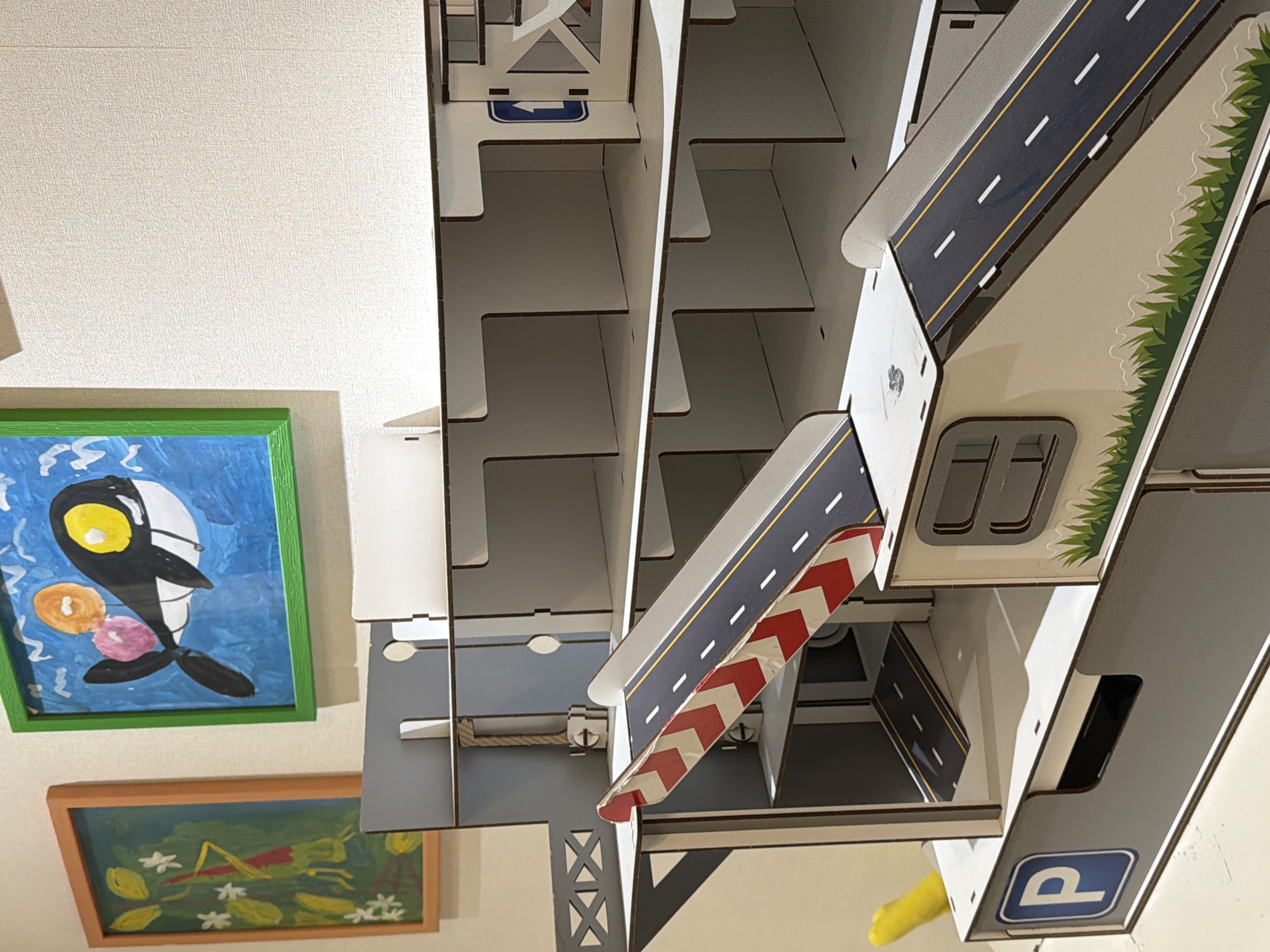 This media file presents an indoor scene featuring a multi-level toy car garage or parking structure, alongside two framed children's drawings displayed on a light-colored, textured wall. The image is taken from an elevated, angled perspective, looking down into the toy structure and across the wall.
The central subject is an elaborate toy garage, constructed from interlocking pieces of what appears to be wood or similar material. It boasts several levels and ramps, some of which are adorned with realistic road markings including white dashed lines and yellow solid lines. A prominent detail is a ramp section with distinct red and white diagonal stripes, suggesting a barrier or warning. The structure also incorporates various signs: a blue square with a white 'P' symbol (parking sign) is visible on a lower section, and another blue square with a white left-pointing arrow is present on an upper level. Some areas within the garage are designed as open bays or shelves. Green, textured patterns on the sides of some ramps simulate grassy verges.
To the left, two framed pieces of children's artwork hang on the wall. The upper drawing, set in a bright green frame, depicts a stylized black and white creature with a large yellow eye and additional orange and pink shapes, against a blue background with white accents. Below it, in a light brown frame, is a drawing of a green grasshopper or insect among green leaves and white flowers, set against a darker green backdrop.
A small, curved yellow object, possibly a soft toy or a piece of fabric, is partially visible in the lower right corner of the frame. The wall exhibits a subtle texture, consistent with wallpaper. The scene captures a static arrangement of toys and artwork, characteristic of a domestic play area in Sofiivska Borshchahivka, Ukraine. No specific activity is occurring, and the time of day is not discernible from the ambient indoor lighting.