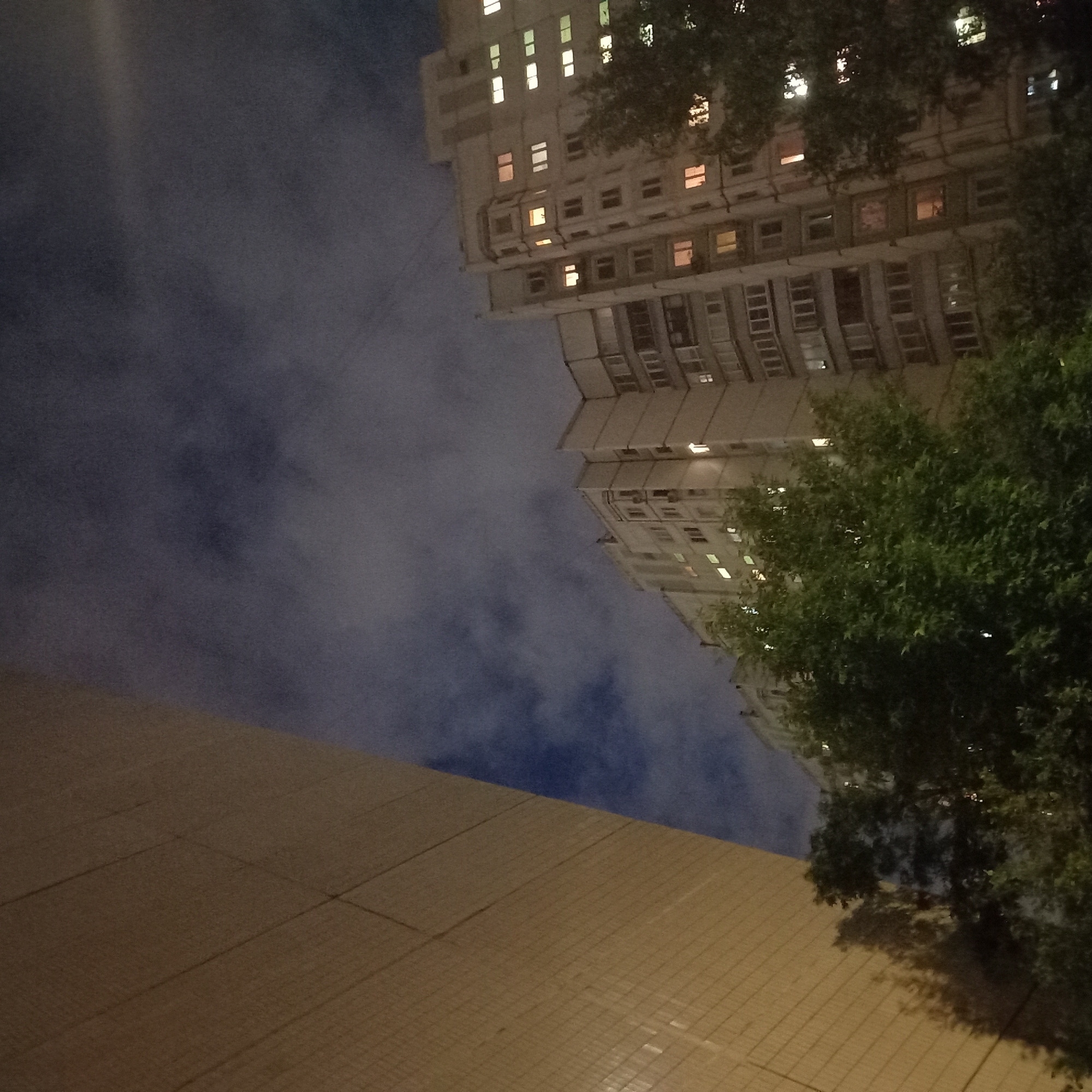 The image is taken at night and shows a low-angle view of a multi-story building and what appears to be a low building next to it. The buildings are located in Moscow, Russia.
The taller building is lit by numerous windows with lights on inside. The lower building has a tiled exterior. In the top right of the image, there is a tree. The sky is dark with some clouds.