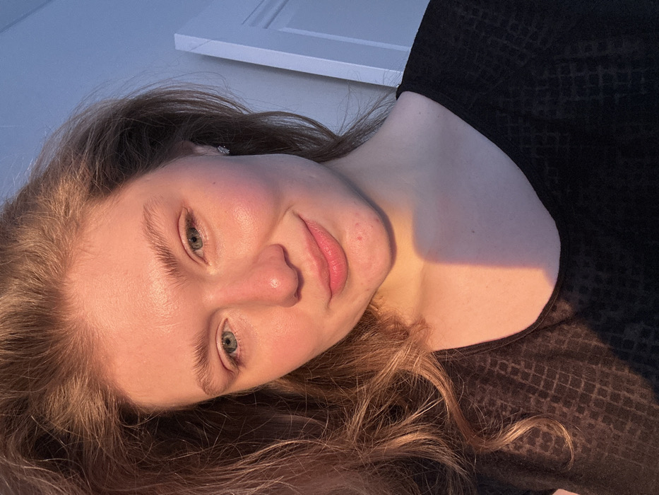The image is a close-up portrait of one young adult female, positioned indoors. The requested location of Kazan, Russia, cannot be determined from the visual information presented in the image.
The subject has light skin, light brown to auburn hair, and blue eyes, which are directed forward towards the camera. She displays a slight, closed-mouth smile. Small blemishes are visible on her chin and jawline. A silver-colored stud earring is present in her right ear. She is wearing a dark, likely black, short-sleeved top with a subtle, textured grid pattern.
Strong, warm, directional light illuminates the left side of her face, hair, and neck, suggesting late afternoon or early evening sunlight entering a window. This light source casts a distinct shadow diagonally across her neck and upper chest. The right side of her face is less intensely lit. The background is softly blurred, showing a light blue surface behind her head and upper body. To the far right, a vertical white structure, consistent with a door frame or wall paneling, is partially visible.