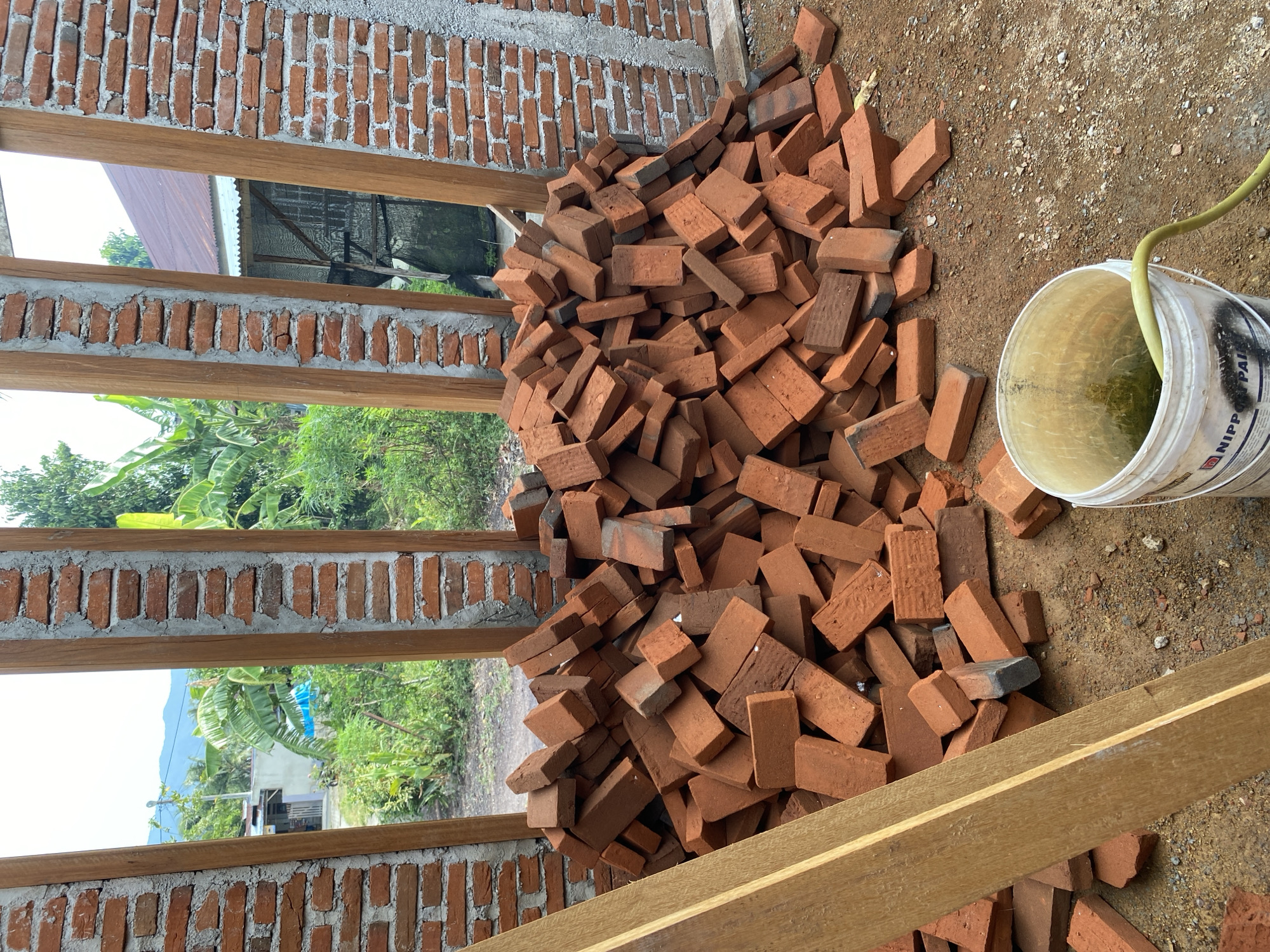 A pile of red bricks is located on the ground in front of a partially constructed brick wall. Wooden beams frame openings in the wall, revealing a view of lush green foliage and distant hills. A white bucket with a yellow hose and murky liquid sits in the foreground to the right of the bricks. The ground is covered in dirt and small pebbles. The overall scene suggests an ongoing construction project in a rural or suburban outdoor setting.
