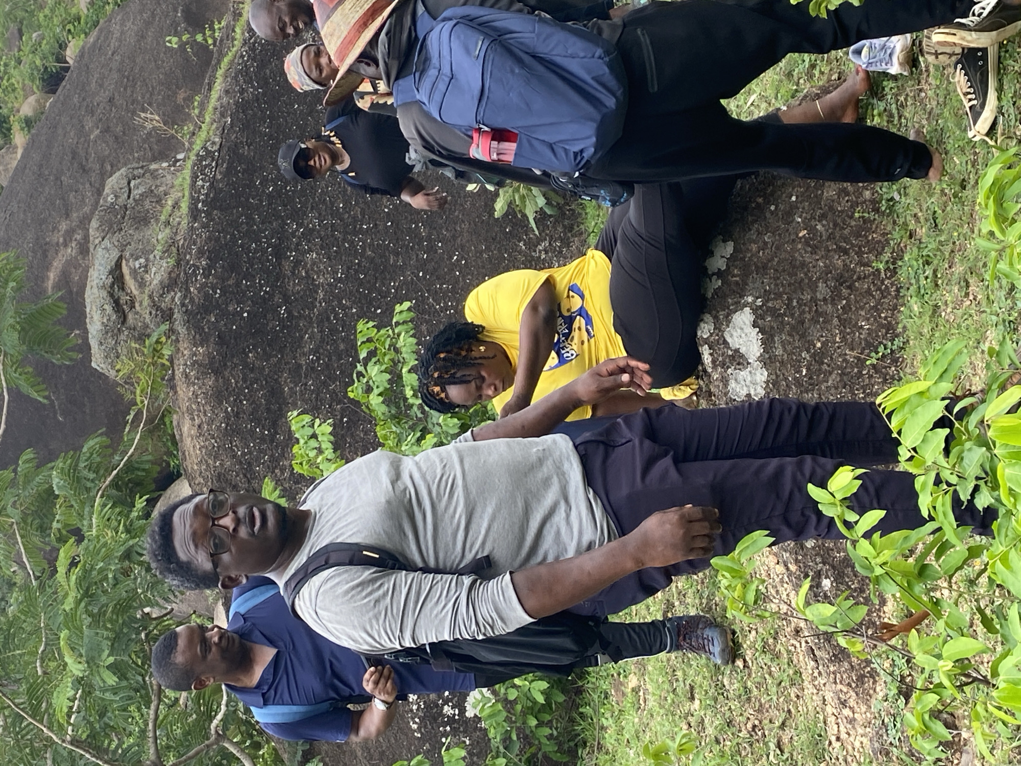 A group of people stand near a large rock formation, likely at the Ri-Yom Rock Formation in Ganawuri, Nigeria. Some of the people are wearing backpacks and hiking shoes. There are bushes and trees nearby. One person is wearing a hat.
