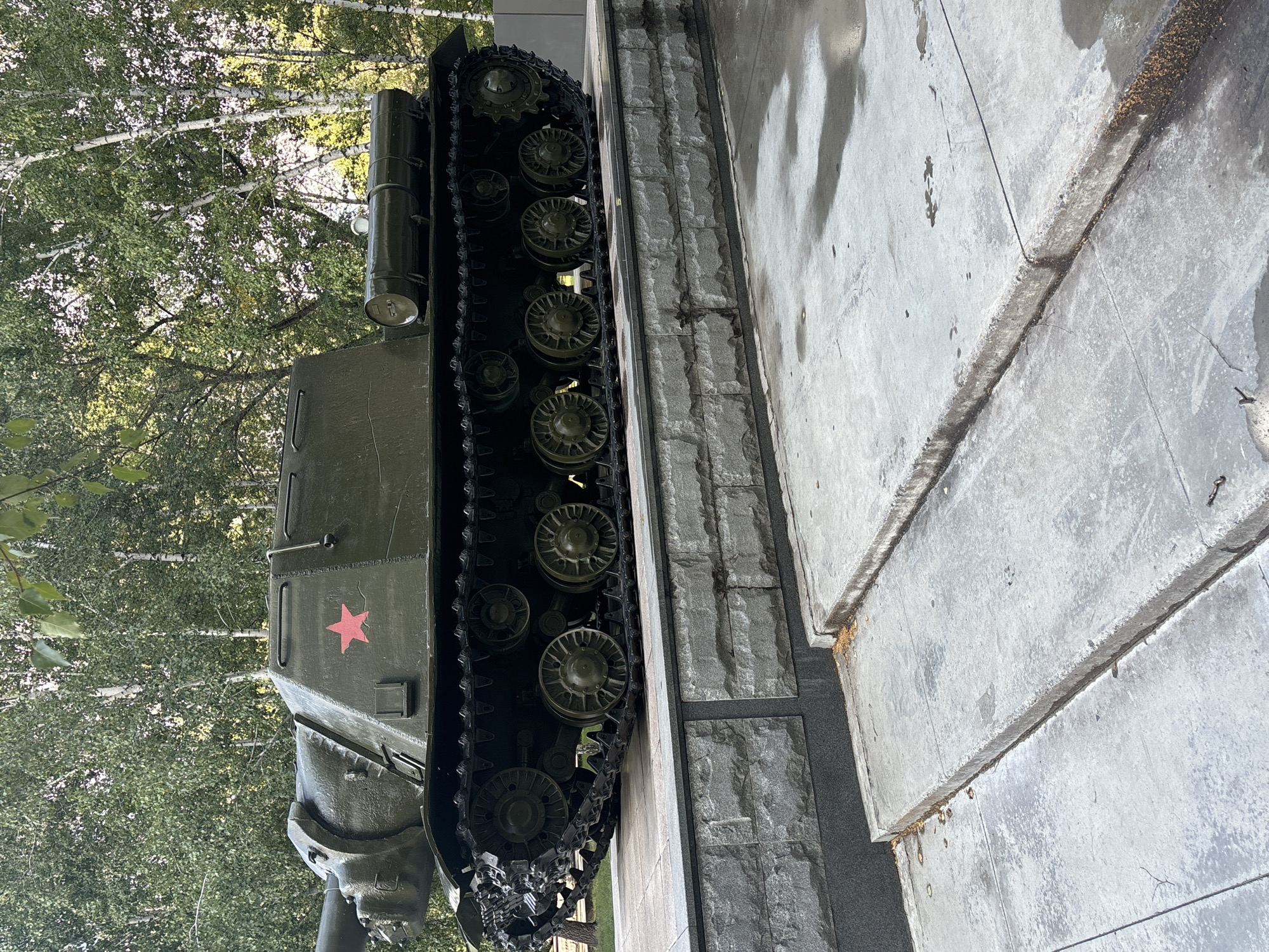 The image depicts a static, outdoor display of a green military tank with a red star insignia, situated on a stone platform in Novosibirsk, Russia. The tank is positioned centrally, its tracks and hull clearly visible. Behind the tank, a backdrop of lush green trees and slender white birch trees suggests a park or memorial setting. The foreground shows concrete steps and a paved area, indicating a public space designed for viewing the monument. The overall scene is one of stillness and remembrance, with no people present.