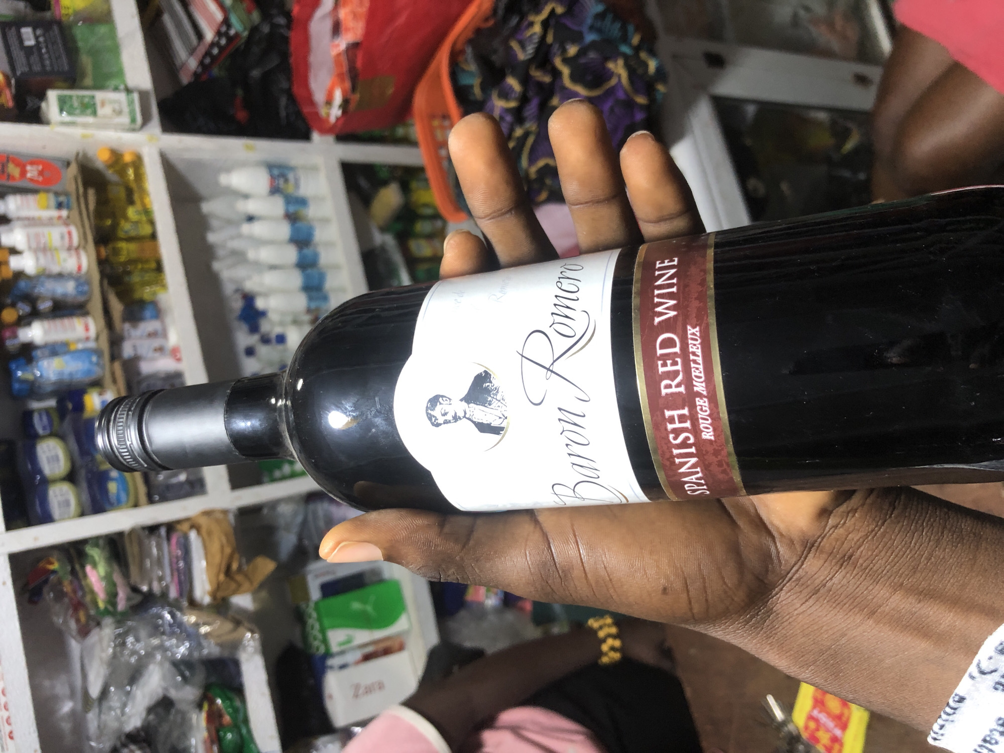 In Ado, Nigeria, a person's hand holds a bottle of Baron Romero Spanish Red Wine. The bottle has a black neck and cap, and a white label with the words "Baron Romero" in elegant script, a portrait of a man in a suit, and "SPANISH RED WINE ROUGE MEILLEUX" in red text. The background shows shelves stocked with various items, including bottles, containers, and bags, suggesting a retail or storage environment. The person is wearing a yellow beaded bracelet on their left wrist.