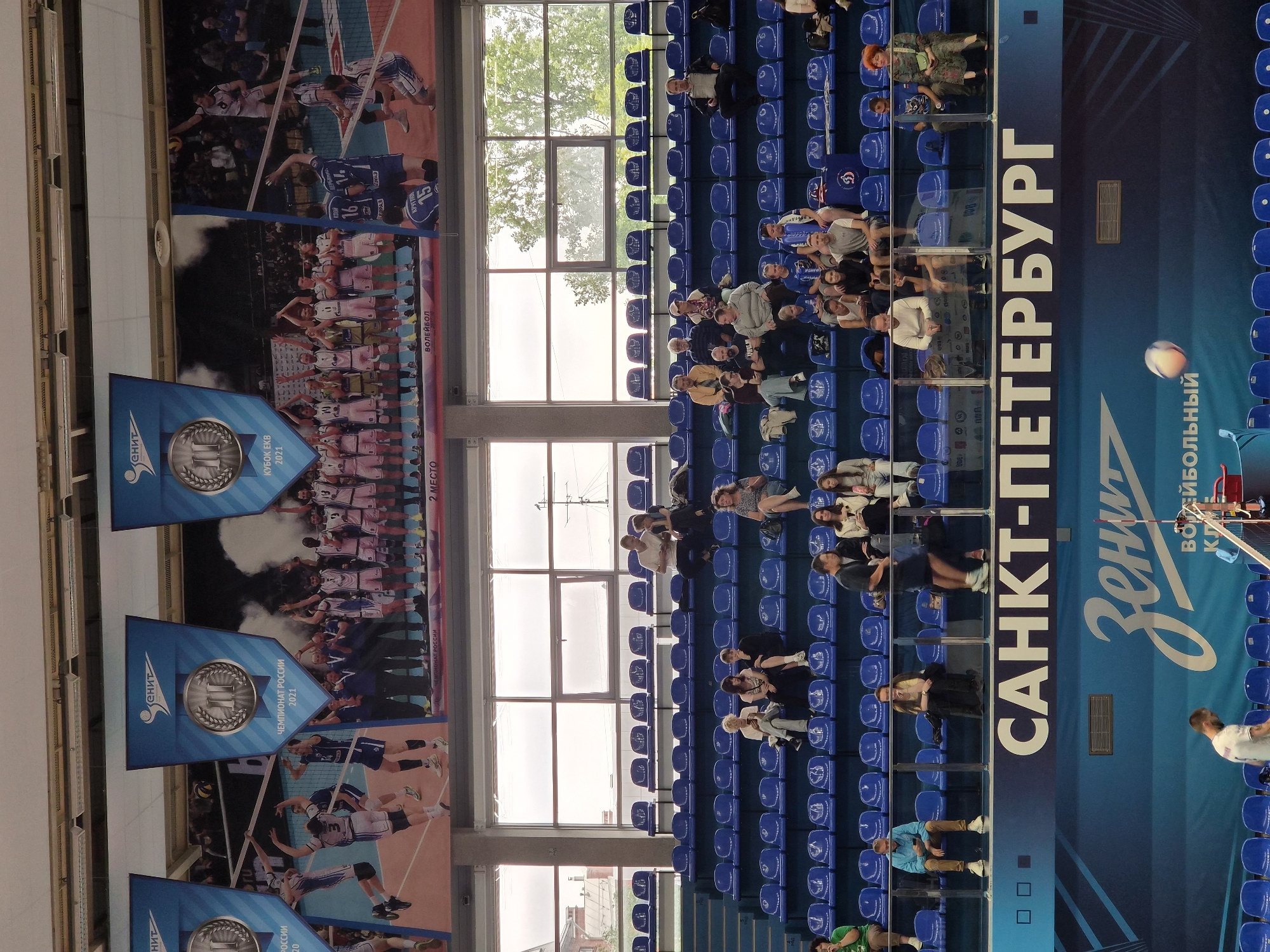 The image depicts the interior of a volleyball arena in Saint Petersburg, Russia. Large banners featuring volleyball team logos and championship graphics hang from the ceiling above the seating areas. The arena features rows of blue stadium seats, some of which are occupied by spectators. The spectators are of various ages and are dressed in casual attire. Some are watching the court, while others are engaged in conversation or using their phones. In the foreground, a volleyball is in mid-air, suggesting an ongoing match. The overall scene conveys a sense of athletic competition and community engagement within the sports venue.