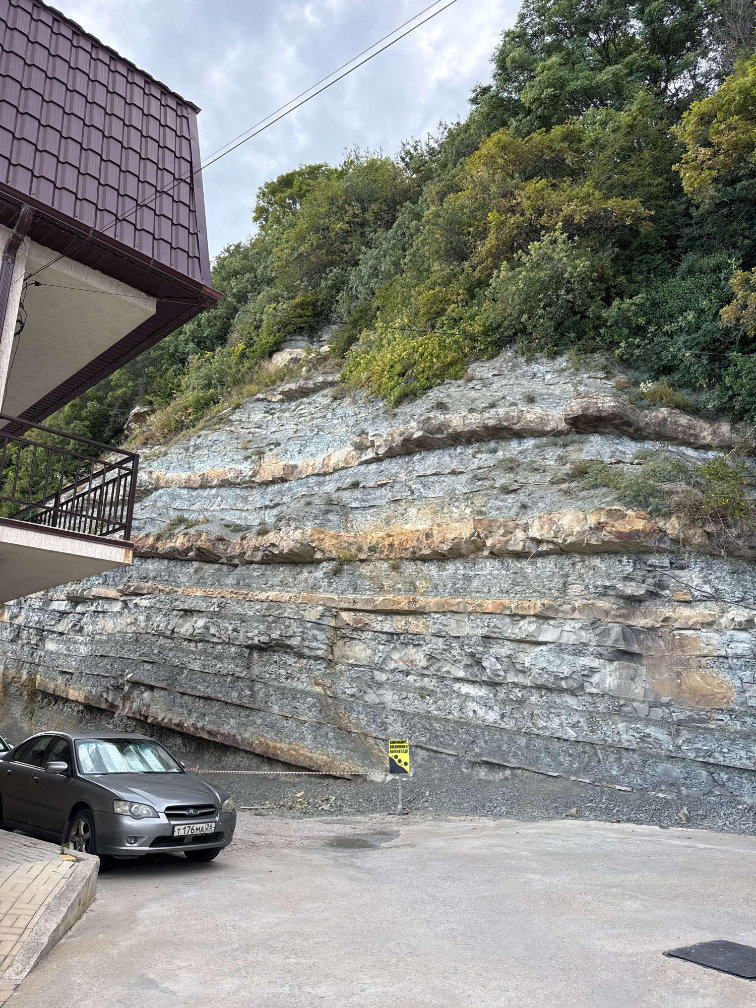 The image depicts a residential building with a brown tiled roof adjacent to a steep, layered rock face. Lush green trees and foliage cover the upper portion of the rock face. A gray sedan is parked on a paved surface in front of the rock face. A yellow warning sign with black text is visible near the base of the rock face. The scene appears to be located in Novomikhaylovsky, Russia, given the presence of a Russian license plate on the car. The overall environment is natural, with the rock formation indicating a geological feature. There are no people visible in the image, and no specific actions or interactions are taking place.