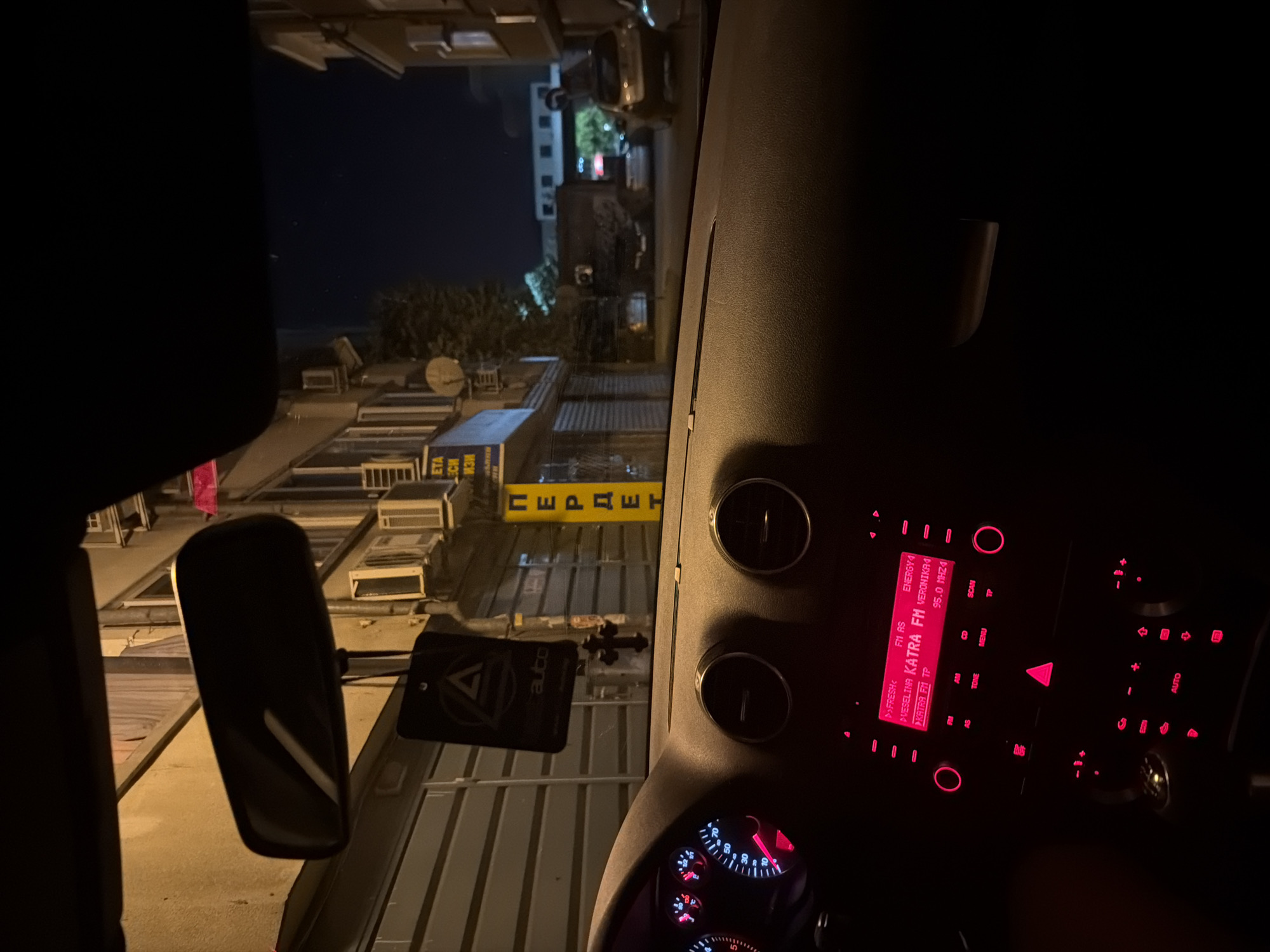 The image shows the interior of a car at night, looking out the windshield. The car's dashboard is visible in the foreground, with its red illuminated radio display and instrument cluster. Outside the car, a street scene unfolds in the city of Plovdiv, Bulgaria. The buildings are lit by streetlights and illuminated signs, one of which reads "ПЕРДЕТ" vertically in yellow letters. The scene depicts a typical urban environment at night, with cars parked along the street.