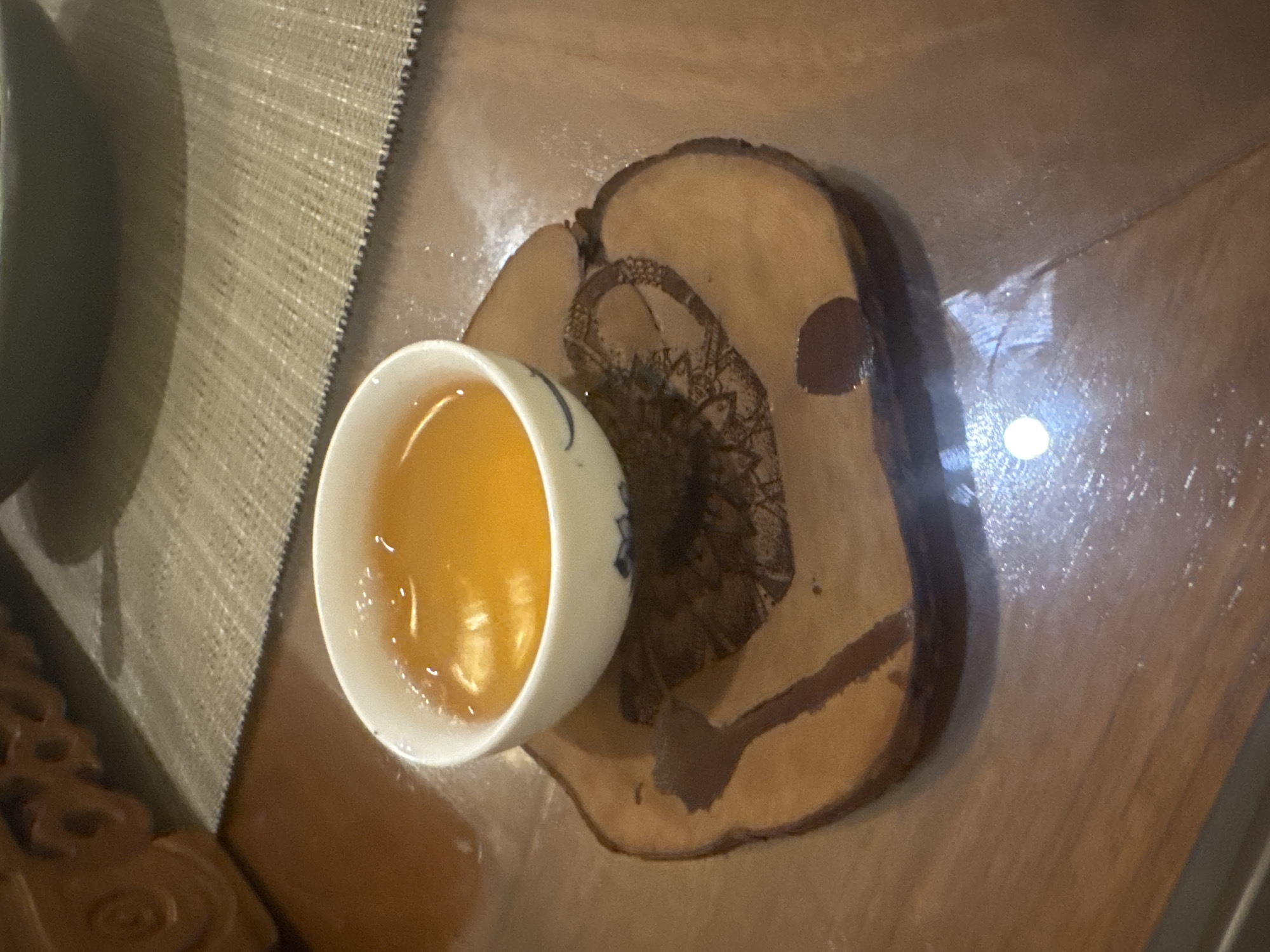 The image shows a teacup filled with a yellow liquid, likely tea, sitting on a wooden coaster with a decorative pattern. The coaster is placed on a brown surface, likely a table. A woven mat or placemat is visible to the left of the teacup. The scene appears to be indoors. There is no information available in the image to confirm this is in Kuta Selatan, Indonesia.