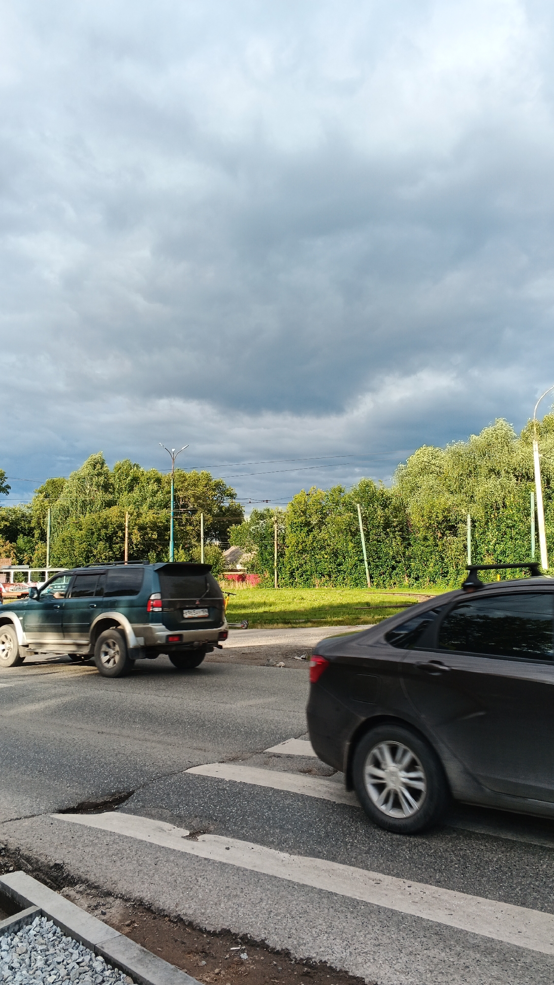 This is a vertical, eye-level, outdoor photograph taken from street level. The scene depicts a road with a crosswalk, two vehicles, and a lush green treeline under a cloudy sky.
In the foreground, a dark-colored sedan is moving across the crosswalk from right to left. The sedan has a roof rack. To the left of the sedan, a dark green SUV is positioned on the road, also moving from right to left. The SUV's rear is visible, with a license plate that appears to be in Russian format. The road surface shows signs of wear and tear, with a notable crack or dip in the crosswalk area. Gravel and dirt are visible on the edge of the road to the lower left.
Behind the road, a dense line of mature green trees forms a natural backdrop, interspersed with utility poles and wires. The sky is filled with heavy, dark grey clouds, suggesting overcast weather. The overall impression is a somewhat dreary day in a suburban or semi-urban environment. The location is identified as Izhevsk, Russia.