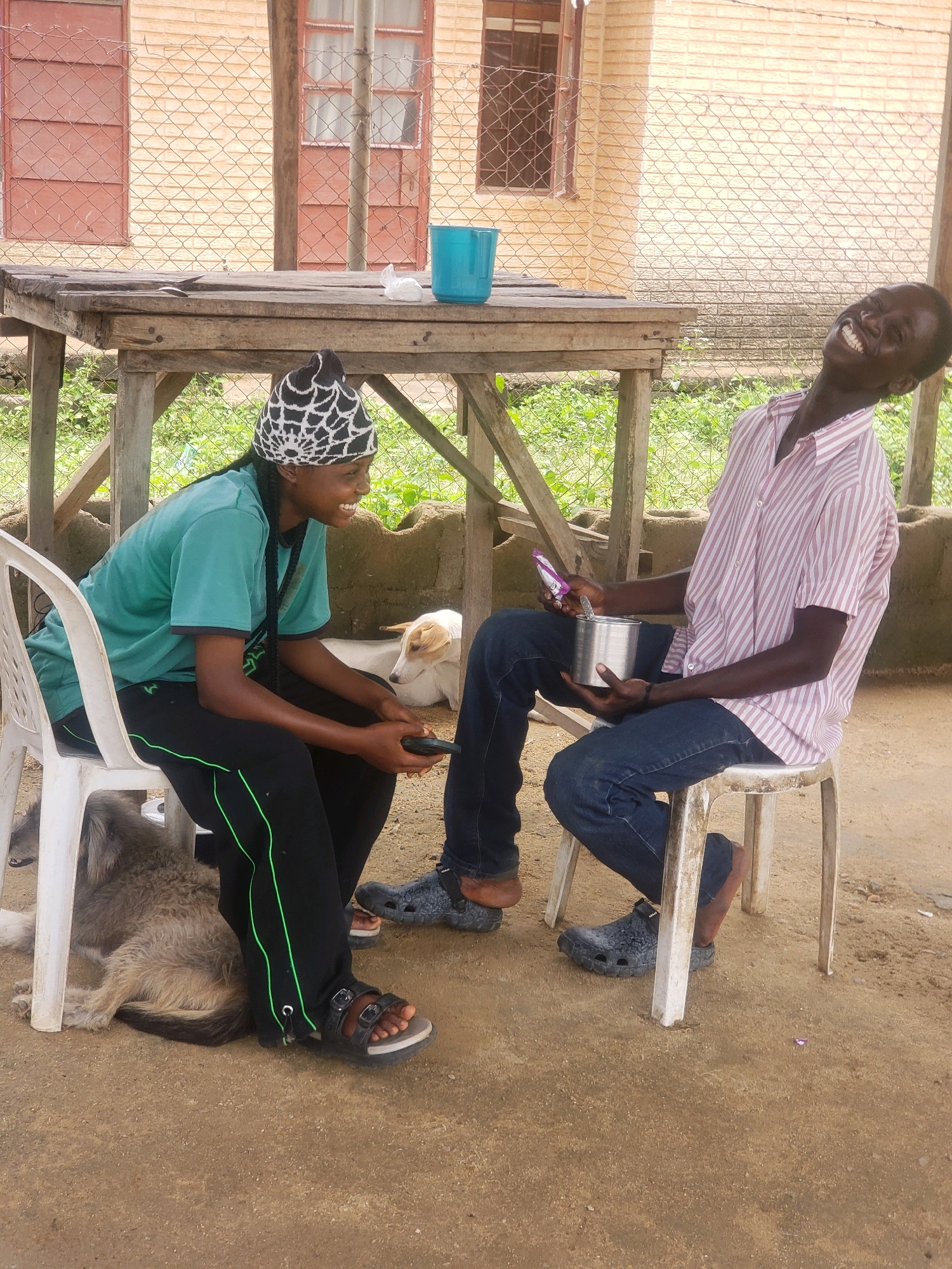 Two individuals are seated on chairs under a wooden structure. The person on the left, wearing a turquoise t-shirt and black pants with green stripes, sits on a white plastic chair and holds a dark object, possibly a phone. They are wearing a black and white patterned beanie and sandals. To their right, on a white stool, sits another person wearing a pink and white striped short-sleeved shirt and blue jeans. This individual is laughing heartily, holding a silver metal container and a small purple packet. The ground is sandy, with some green vegetation visible in the background. Behind the people, a beige brick building with red-framed windows is visible, partially obscured by a wire fence. A dog lies on the ground near the person on the left. The scene appears to be an outdoor seating area. The location is identified as Narayi, Nigeria.