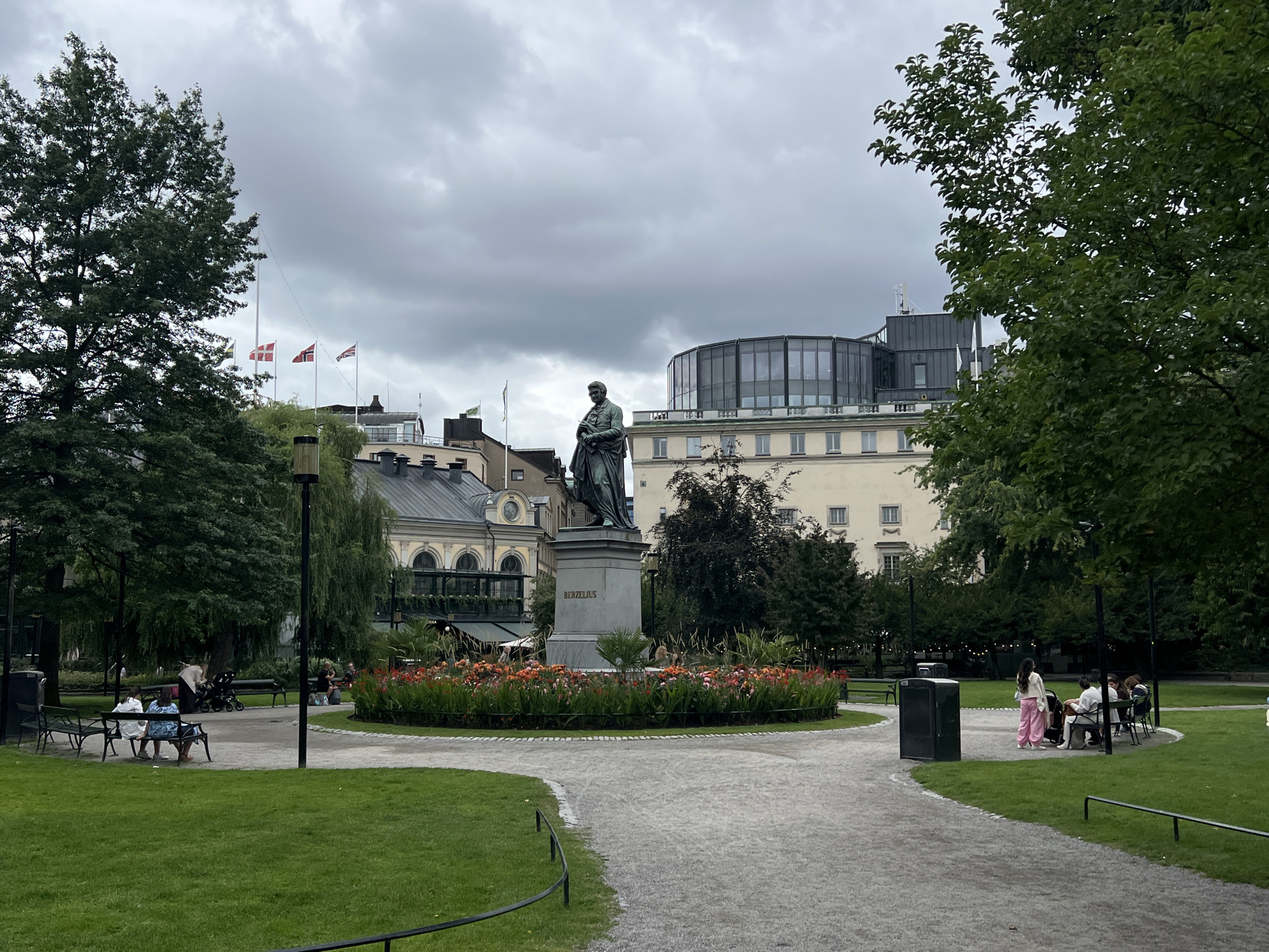 This image depicts the Berzelius Park in Stockholm, Sweden. In the center of the frame, a bronze statue of a man in historical attire stands on a stone pedestal inscribed with "BERZELIUS". The statue is surrounded by a circular bed of vibrant orange and red flowers. A gravel path curves around the flower bed, separating it from a well-maintained lawn.
Several people are visible in the park. On the left side, two women and a child are seated on a bench, facing away from the camera. A stroller is parked behind them. Further to the right, a woman in a white top and pink pants stands near a dark trash receptacle. Several other individuals are seated on benches in the background, appearing to relax or socialize.
Behind the statue and flower bed, buildings with light-colored facades and dark roofs are visible. One building has a distinctive curved glass facade on its upper level. Flags are flying from poles in front of some of the buildings. Lush green trees with dense foliage frame the scene, with one large tree on the left and another on the right side of the image. The sky is overcast with a cloudy, gray appearance. The overall atmosphere is calm and serene, suggesting a public park setting on a cloudy day.