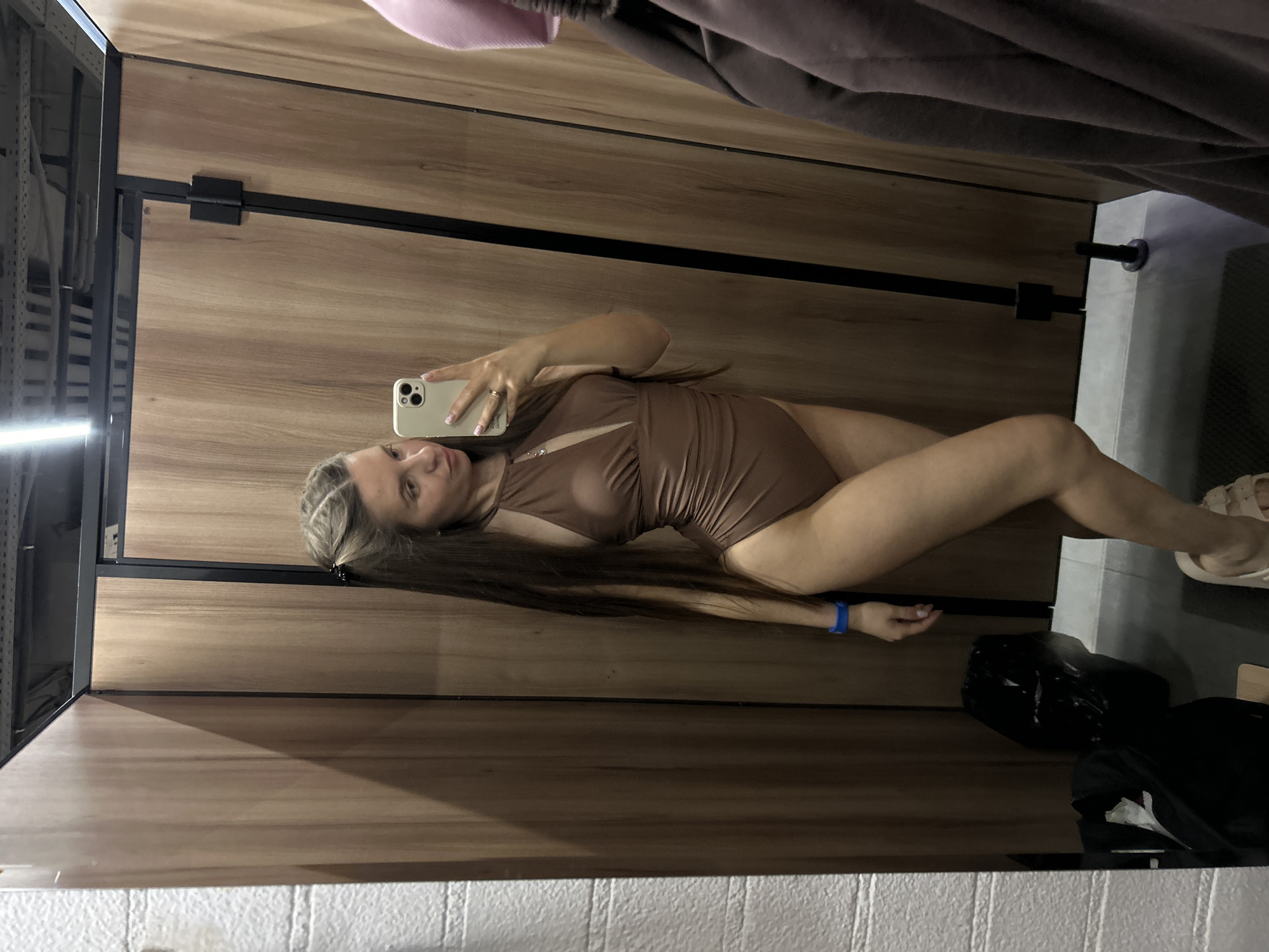 The image displays a woman taking a mirror selfie in an indoor setting, likely a changing or fitting room. The photograph is oriented ninety degrees clockwise, showing the woman lying on her left side. She has long, dark hair and is wearing a brown or dark beige one-piece swimsuit or bodysuit featuring a cutout detail on the chest. She holds a light-colored smartphone, possibly an iPhone, in her right hand to capture the picture. A ring is visible on her right ring finger, and a blue wristband is on her left wrist. She is also wearing light-colored sandals on her feet.
The background features vertical wooden wall panels with visible black metal framing and hinges, suggesting a partition or door within the changing area. A bright light source is reflected on the left side of the mirror. In the upper part of the image, dark fabric, possibly clothes, is visible. On the floor in the lower right corner, a black plastic bag and another dark bag are present. The very bottom edge of the frame reveals a white, textured surface, resembling a tiled floor or a lower wall section. The woman has a neutral expression, looking directly at the phone's camera. Based on the provided context, the location is likely in Tver, Russia. No visible text is present in the image.