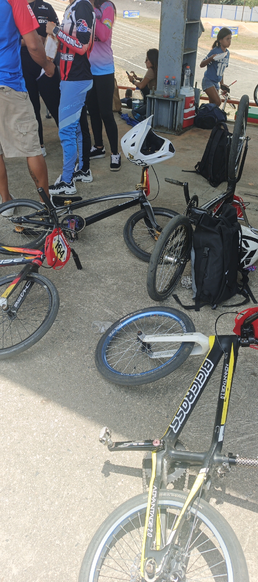 Here is a forensic description of the image:
**Location:** The image seems to be in Tinaquillo, Venezuela, at a BMX track or similar race facility.
**Scene:** The foreground is filled with several BMX bikes laid on the concrete ground. A black BMX bike has a helmet on the handle, and another bike is partially visible with the words "BICICROSS ADVANTAGE 2.0" on its frame. There is a red helmet with the number "164" attached to the handlebars of one bike. Backpacks are nearby, suggesting the cyclists are likely in a waiting or resting area.
**People:** Several people are in the background. They appear to be race participants, spectators, or staff. Some are standing and conversing, while others are sitting or observing the scene. A young girl stands near the edge of the concrete platform, possibly eating food from a plate. Other people are taking pictures.
**Objects:** Besides the bikes, helmets, and backpacks, there are water bottles, a cooler, and race track elements visible in the background.