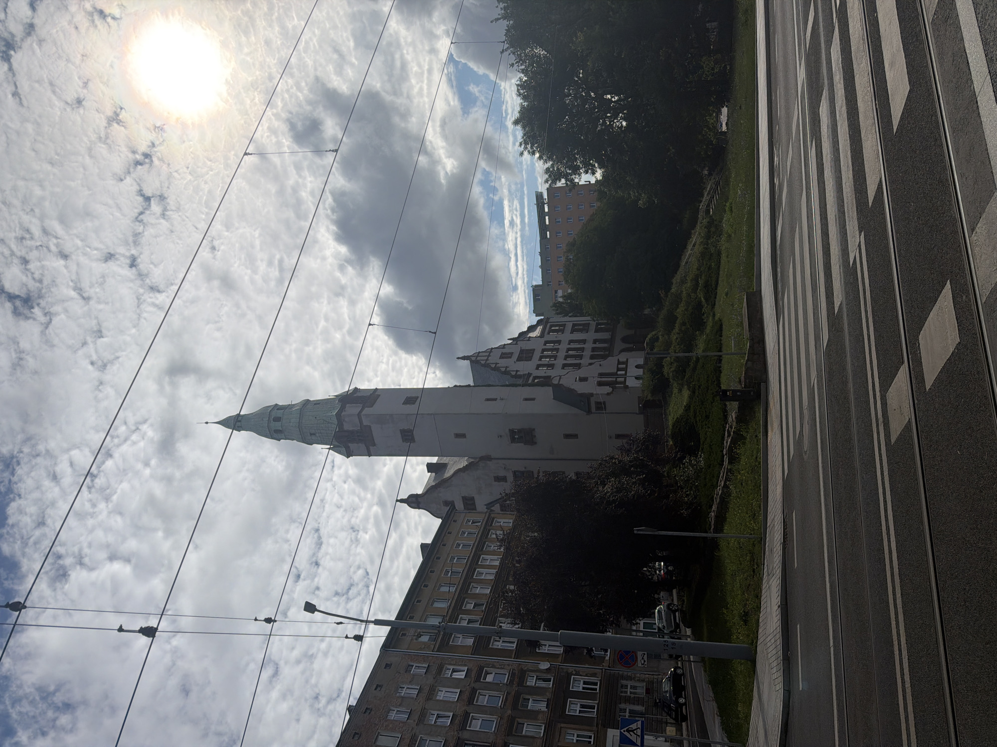The image shows a view of a street in Szczecin, Poland, with a prominent building featuring a tall, green-spired tower. The building appears to be historic, with several sections and different architectural styles. Adjacent to it is a multi-story residential building. In the foreground, there is a street with tram tracks and a pedestrian crossing. The sky is partly cloudy, with the sun visible and casting a bright halo. Several overhead wires, likely for the tram system, cross the frame. There are a few cars visible on the street. The overall impression is a typical urban scene on a cloudy day.