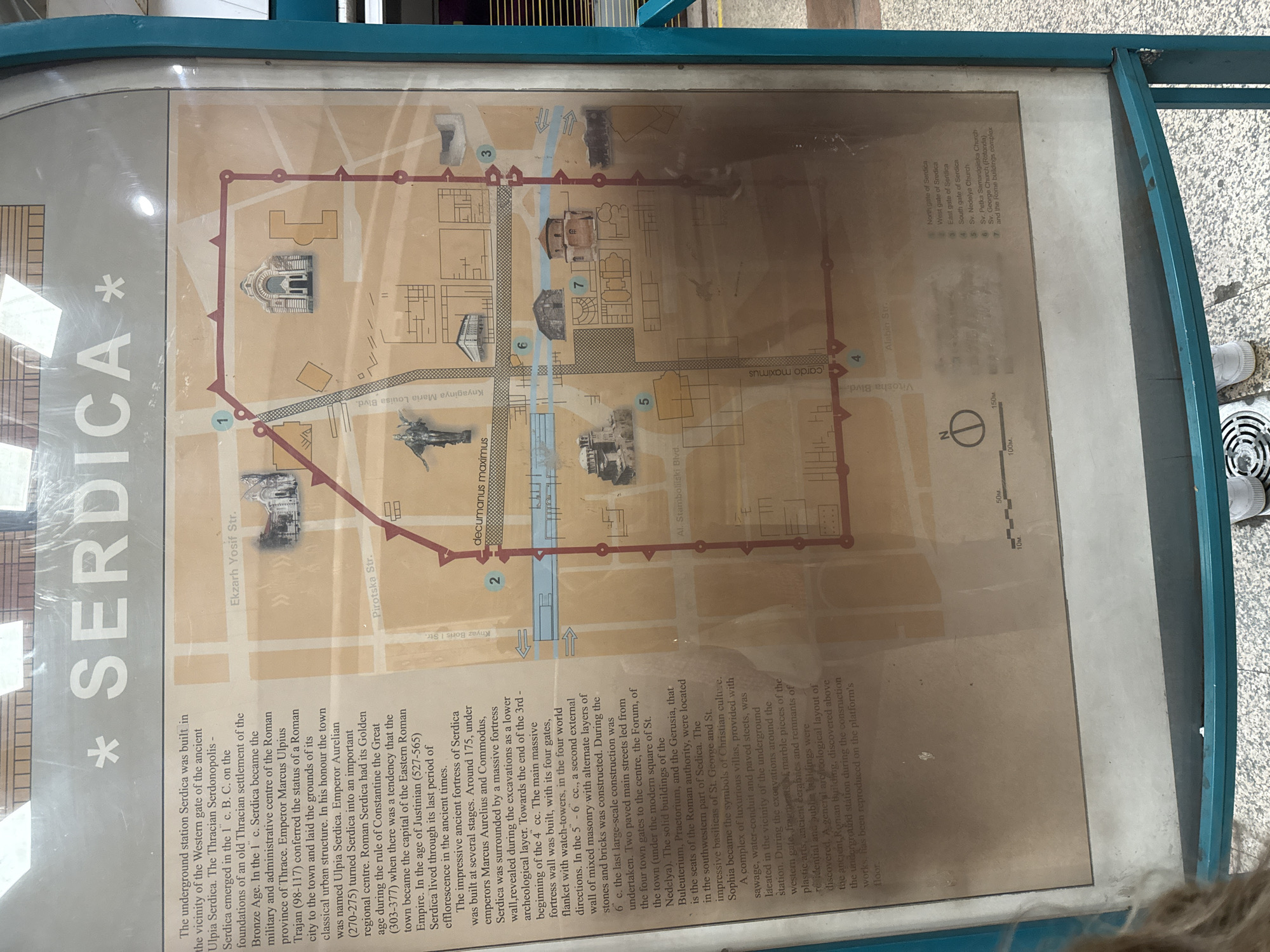 This image shows a map of ancient Serdica (now Sofia, Bulgaria) displayed on a sign near the Serdica underground station. The map outlines the boundaries of the Roman-era city with a red line, marking its gates in the north, west, east, and south (numbered 1-4). Major thoroughfares like "decumanus maximus" and "cardo maximus" are labeled. Key buildings such as the churches of Sveta Nedelya, Sveta Petka Samardjiyska, and Saint George Rotunda are indicated, along with the Roman buildings complex. The map includes a scale, providing dimensions in meters. The text to the left details the history of Serdica, from its Thracian origins through its Roman golden age and subsequent periods.