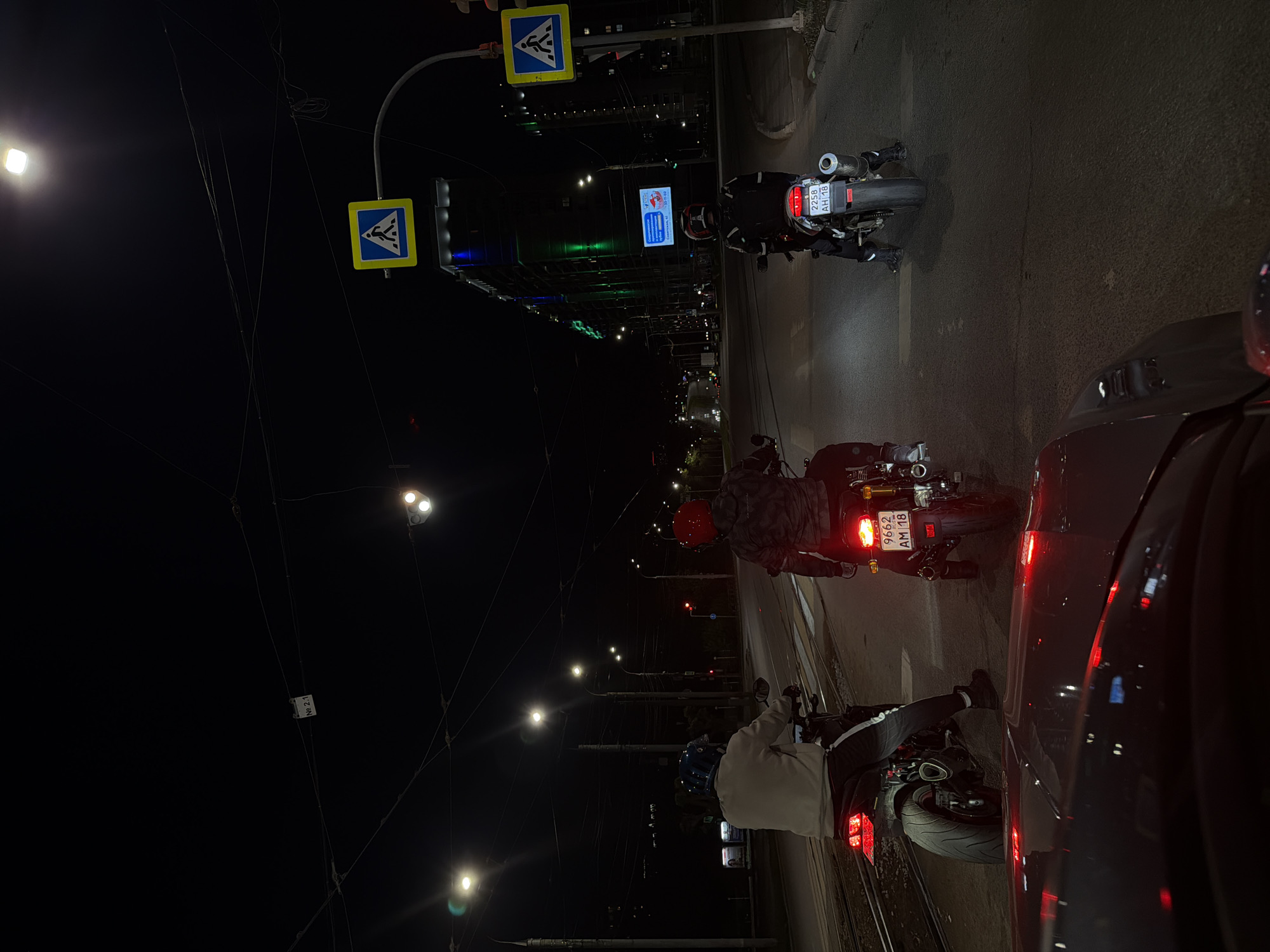 The image depicts a street scene at night in Izhevsk, Russia. Several motorcycles are stopped at an intersection, illuminated by streetlights. Pedestrian crossing signs are visible overhead. A tall building with green illumination is in the background. Cars and motorcycles are stopped in traffic. License plates on some motorcycles are visible.