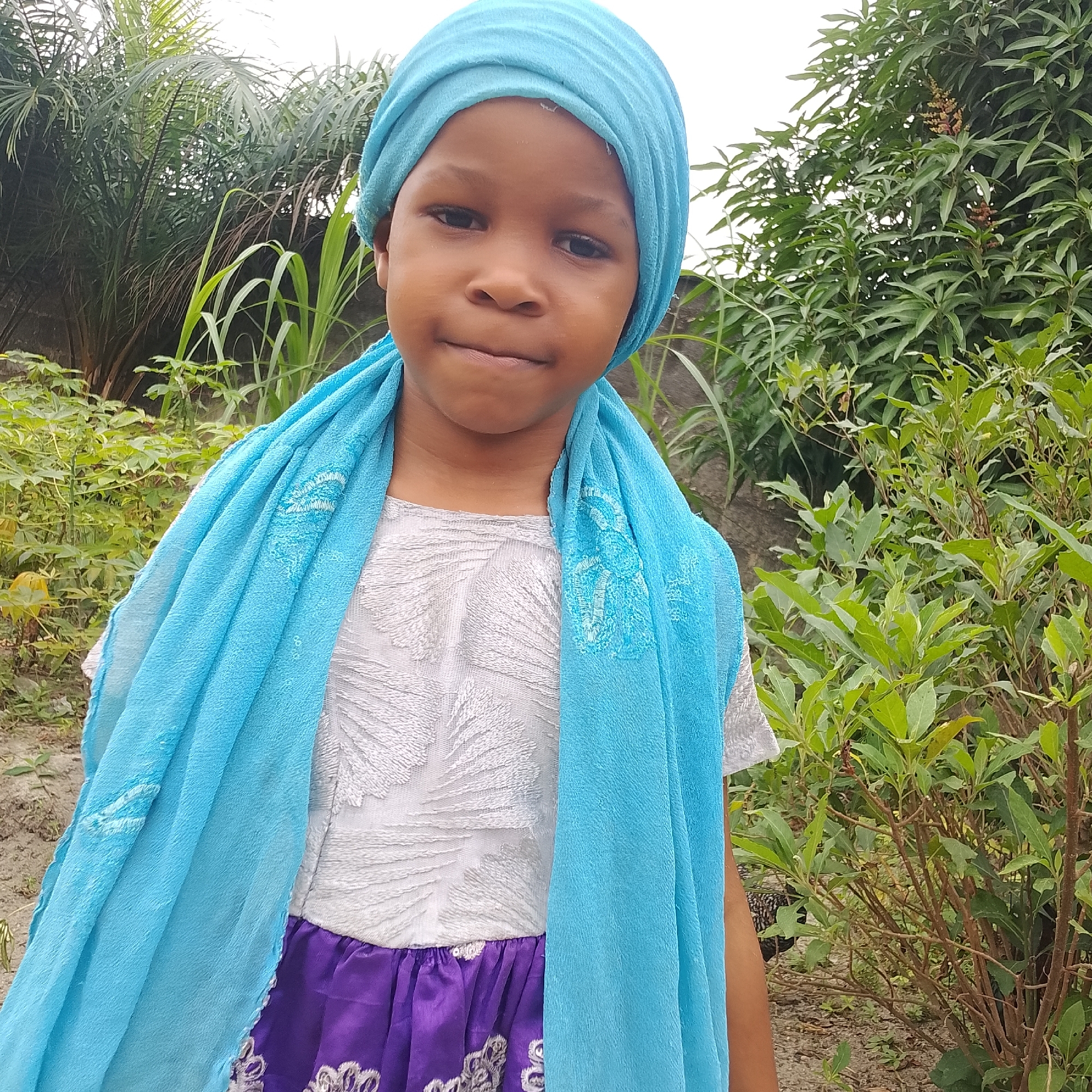 In Ado, Nigeria, a young girl stands outdoors, positioned slightly off-center to the right. She is facing the camera, her gaze directed forward. The girl is wearing a blue headscarf and a light blue scarf draped around her shoulders. Her attire includes a white top with intricate detailing and a purple skirt. The background consists of lush green foliage, including palm trees and other plants, with a neutral-toned wall visible behind her. The ground appears to be covered in dirt and small plants. The lighting suggests it is daytime, with an overcast sky.