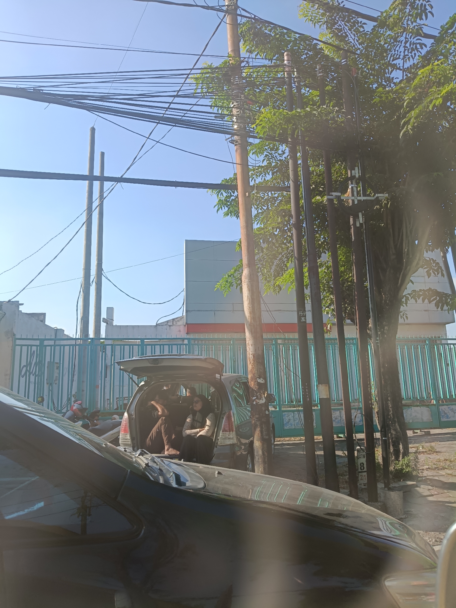 In Klangonan, Indonesia, a scene unfolds on a sunny day under a clear blue sky, with numerous electrical wires strung across the frame. In the foreground, the hood and windshield of a dark car are visible, reflecting the environment. Behind it, a silver hatchback SUV has its rear liftgate open, revealing three individuals. Two people are seated in the back of the vehicle, with one wearing glasses and a striped shirt, and the other in brown trousers. A third person appears to be lying down in the rear of the car. A light blue metal fence runs along the background, behind which is a building with grey and orange accents. Several utility poles, some made of wood and others metal, stand amidst the foliage of a tree.