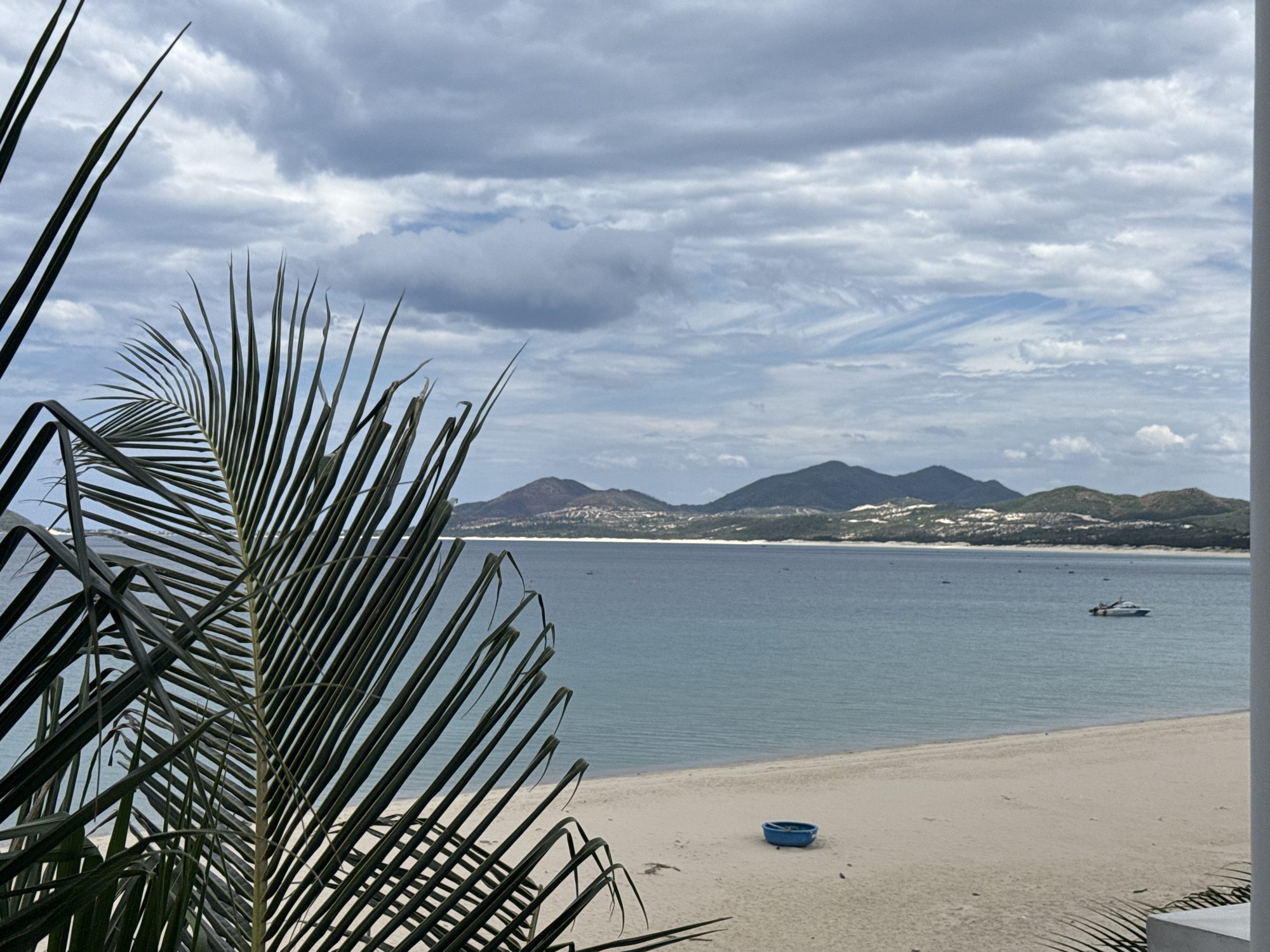 An outdoor, eye-level, wide shot shows a beach scene with palm fronds in the foreground. The beach is made of light sand and is mostly empty, with a small blue boat visible on the sand. The ocean is a calm, turquoise color, and a white and blue motorboat is visible in the distance. The background features a range of green mountains with buildings on their slopes, and a cloudy sky. The overall lighting is subdued, suggesting an overcast day.
