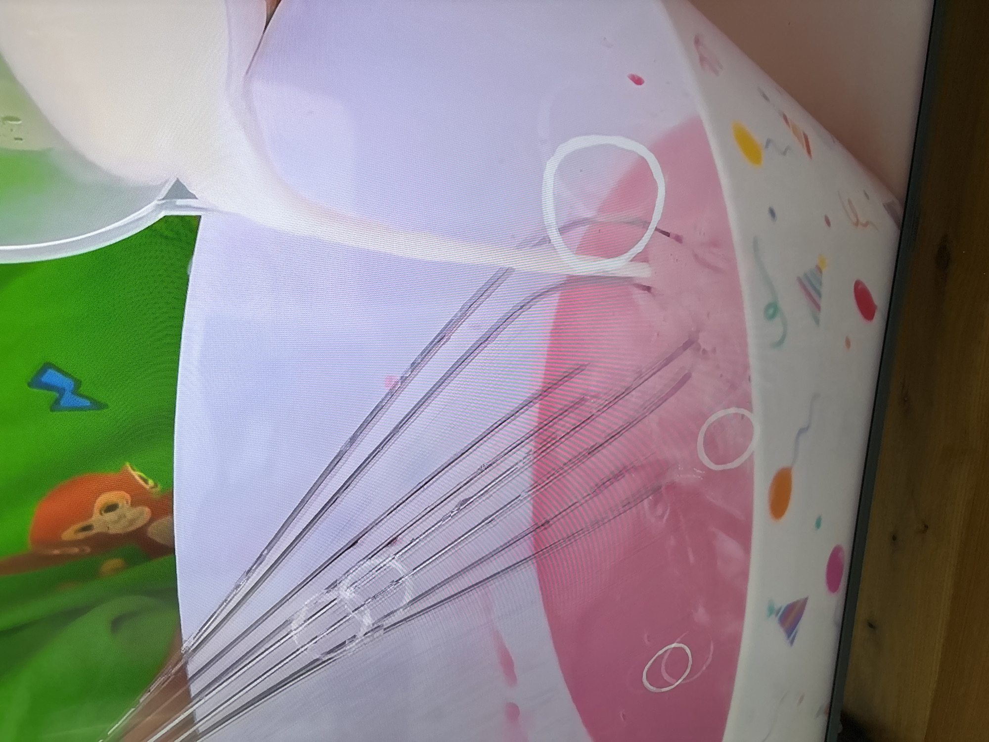 A close-up, eye-level shot captures a pink liquid being poured into a white mixing bowl, which is decorated with a party pattern of balloons and confetti. A metal whisk is partially submerged in the pink liquid. In the upper left corner of the image, a cartoon monkey with orange fur and black eyes is visible against a green background. The scene appears to be indoors, possibly in a kitchen, with a wooden surface visible at the bottom of the frame. There are no discernible landmarks or indications of the city of Dubai, United Arab Emirates, in this image.