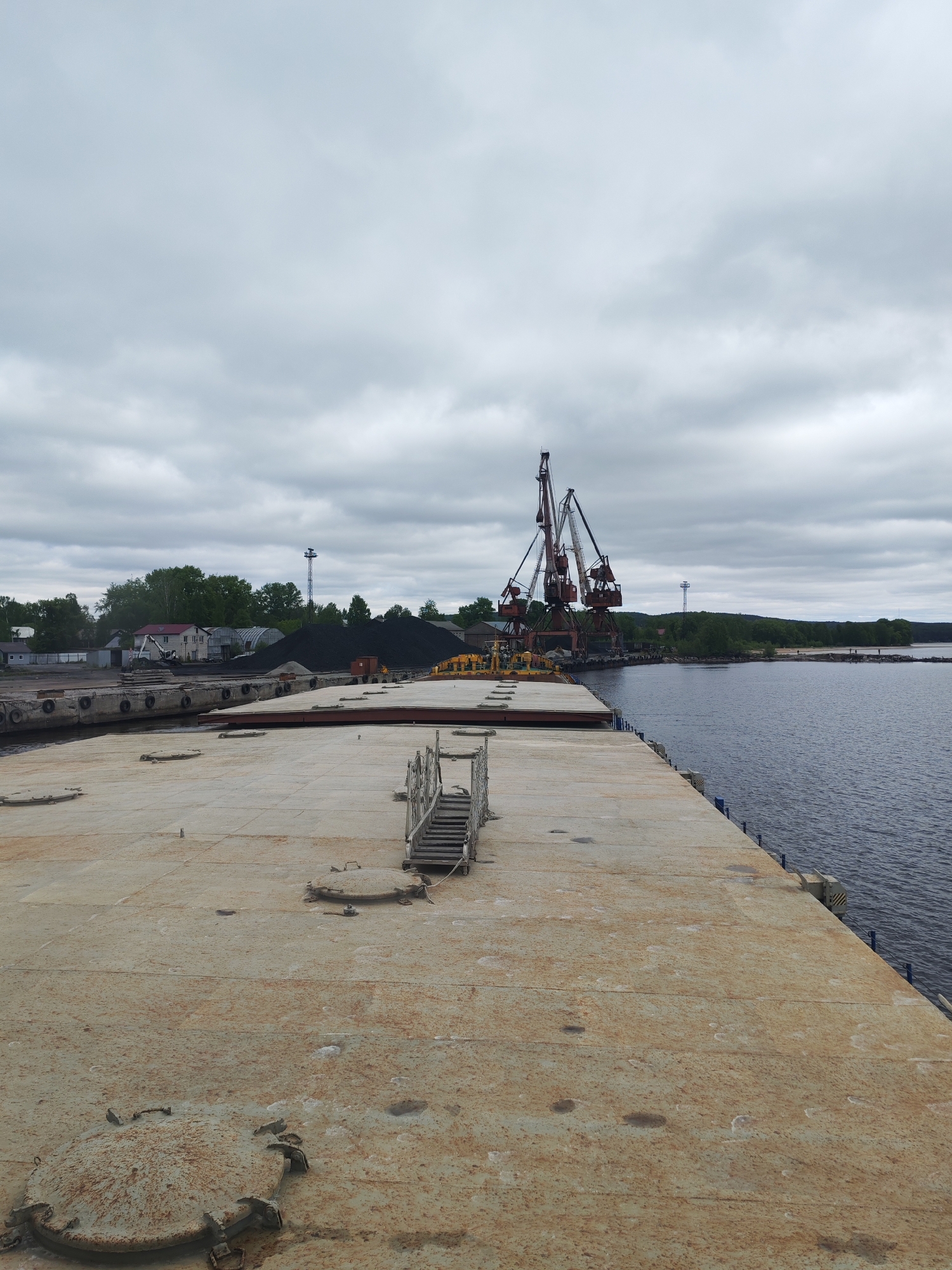 The image presents an industrial port scene under an overcast sky in Medvezhyegorsk, Russia.
In the immediate foreground, a wide, flat deck surface occupies the majority of the lower half of the frame. This surface appears to be made of concrete or metal, heavily weathered and stained with rust, and features several circular metal hatches. A small, rustic set of wooden stairs or a gangway is positioned on the right side of this deck, suggesting it is part of a large barge or pier.
Beyond this foreground structure, to the left, lies the shoreline. Several buildings, including a white house with a red roof, are nestled amongst green trees. Prominent piles of black material, characteristic of coal, are stored on the land, indicating a bulk cargo facility. A tall, slender mast or light tower is also visible amongst the foliage.
In the midground, dominating the center-right, are two large, rust-colored industrial cranes with complex lattice structures. These cranes are situated on a pier or dock, adjacent to a calm, dark body of water, which extends into the background on the right. The presence of cranes and coal piles points to active port operations, likely involving the loading or unloading of cargo.
The background shows a distant treeline bordering the water, with a narrow strip of sandy or rocky shore visible. The sky is entirely overcast, filled with grey clouds, creating soft, diffused light across the scene. No direct sunlight is apparent. The overall impression is one of a functional, industrial environment. No visible text or people are explicitly identifiable in the image, though the infrastructure clearly implies human activity.