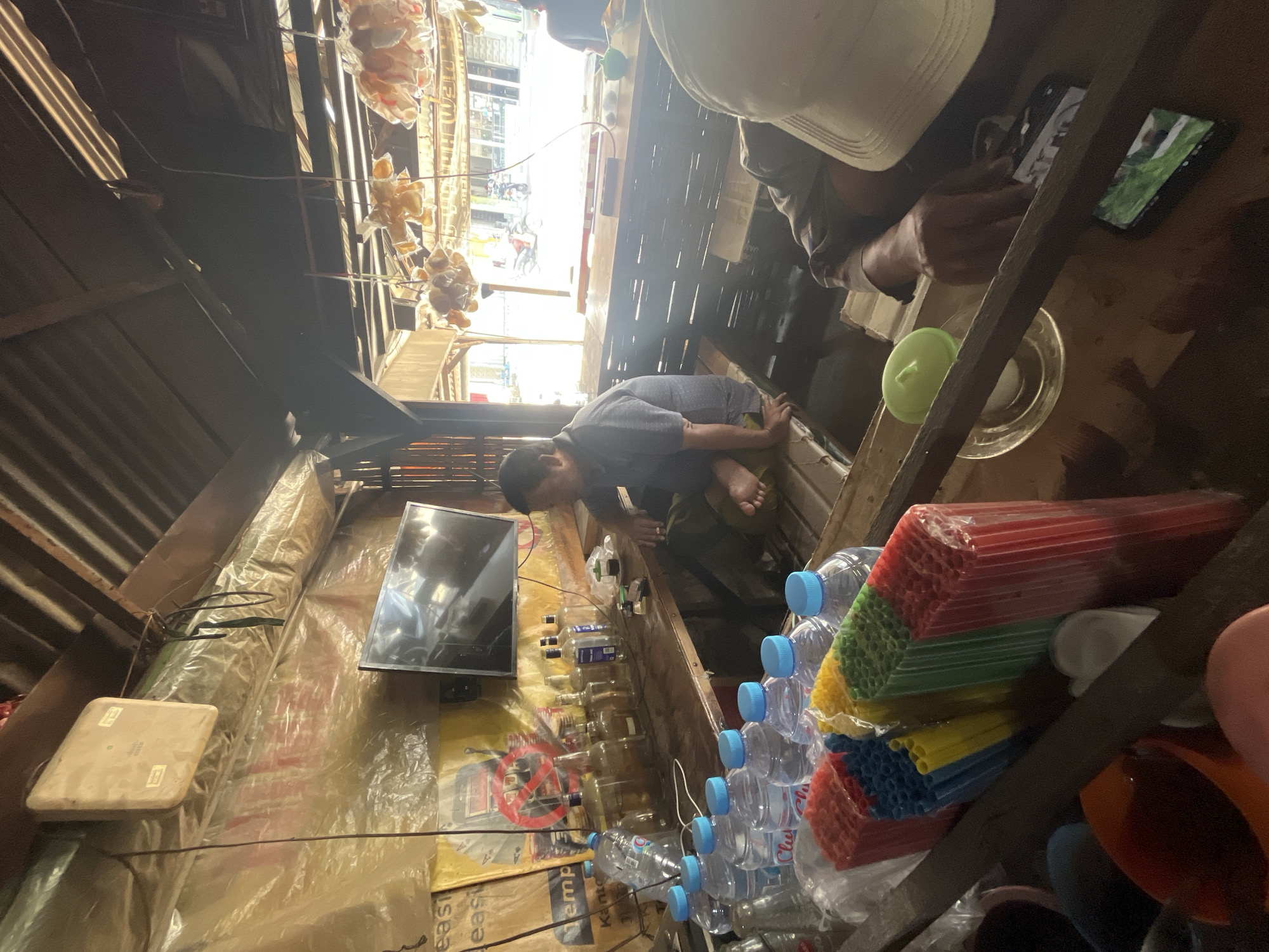 The image depicts an interior scene of a roadside stall in Dusun Metatu, Indonesia. A man, wearing a blue and grey checkered shirt and dark pants, is seated on a wooden platform, facing away from the camera and towards the right. He is holding what appears to be a cigarette and has his right hand resting on his knee. To his left, on the wooden counter, are several empty glass bottles, some with labels, and a small cup with a dark liquid. Above the counter, a flat-screen television is mounted on a wall covered in yellow plastic sheeting.
In the foreground, a collection of plastic water bottles with blue caps is visible, along with stacks of colorful plastic straws, including red, blue, yellow, and green. A person's hand, wearing a beige cap, is visible in the lower right corner of the image, reaching towards the counter. A mobile phone is placed on the counter, displaying an image of green grass.
The stall appears to be open to the street, with daylight illuminating the scene from the right. The ceiling is constructed of corrugated metal. In the background, through the open front of the stall, a street scene is visible with motorcycles and pedestrians. Several items are hanging from the ceiling, possibly food items in plastic packaging. The overall impression is that of a small, informal business operating in a rural or semi-urban environment in Indonesia.