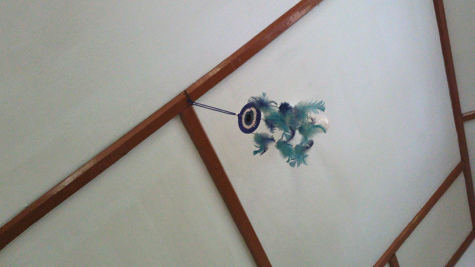 The image shows a room with light blue walls and dark brown wooden beams. An object resembling an evil eye talisman, decorated with blue and green feathers, hangs from one of the beams with a blue cord. The location is in the city of Haini, India. No people are visible.