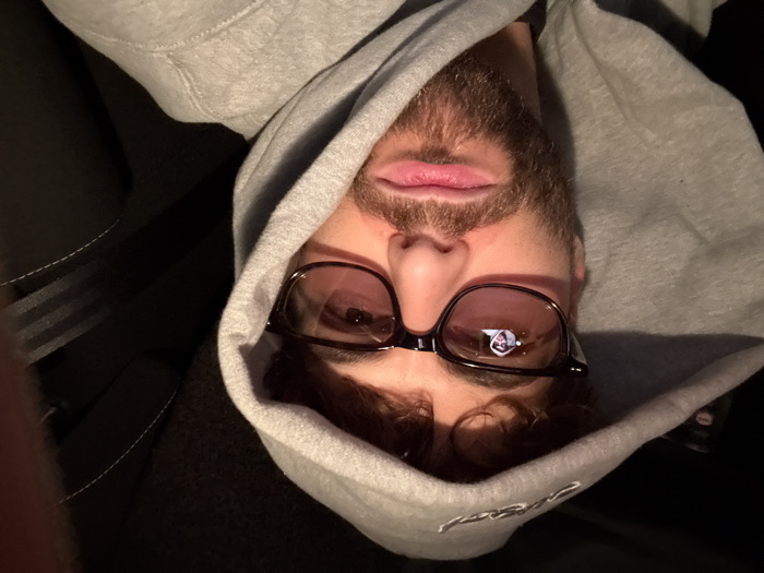 A close-up selfie shows an adult male seated in the interior of a vehicle in Givors, France. The male has light brown, curly hair and a beard. He is wearing a grey hooded sweatshirt with light embroidery on the hood, and dark-framed eyeglasses with lightly tinted lenses. His expression is neutral, with lips slightly pursed. A distinct reflection of a small figure, appearing to be the subject himself in a hoodie, is visible in the left lens of his eyeglasses. The background is dark, indicating a dimly lit environment, possibly night-time. Portions of a dark vehicle seat with white stitching and a seatbelt are discernible on the right side of the frame.