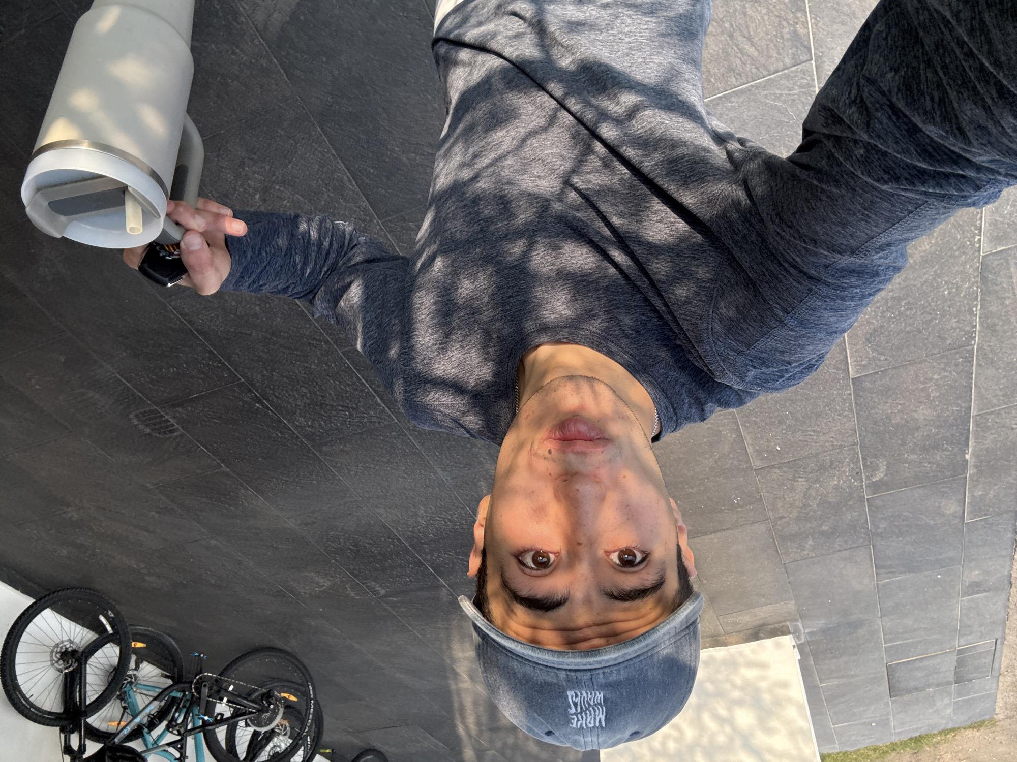 A person, turned upside down, is visible in a selfie. The person is wearing a grey long-sleeved shirt and a blue baseball cap. They are holding a white water bottle in their left hand. A bicycle is partially visible in the lower-left corner of the frame. The setting appears to be outdoors on a tiled surface. The picture was taken in San Bernardino Tlaxcalancingo, Mexico.