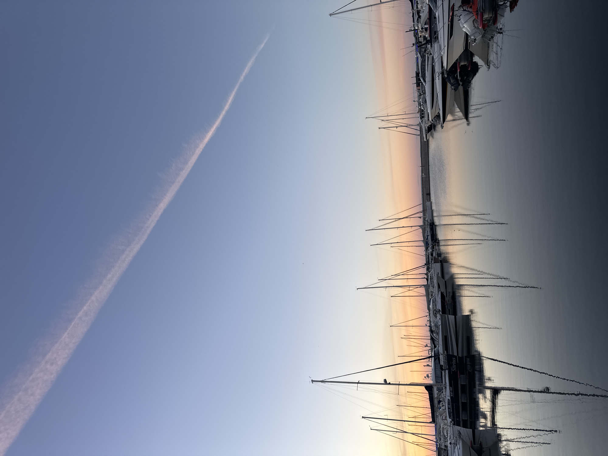 This image depicts a marina at dusk, likely in Tallinn, Estonia, with a prominent contrail stretching across the pale blue sky. Numerous sailboats and motorboats are moored in the calm water, their masts and reflections creating a symmetrical pattern. The horizon displays a gradient of soft orange and pink hues, indicative of the setting sun. The water is still and reflects the sky and the docked vessels. There are no people visible in the frame. The scene suggests a peaceful evening in a coastal urban setting.