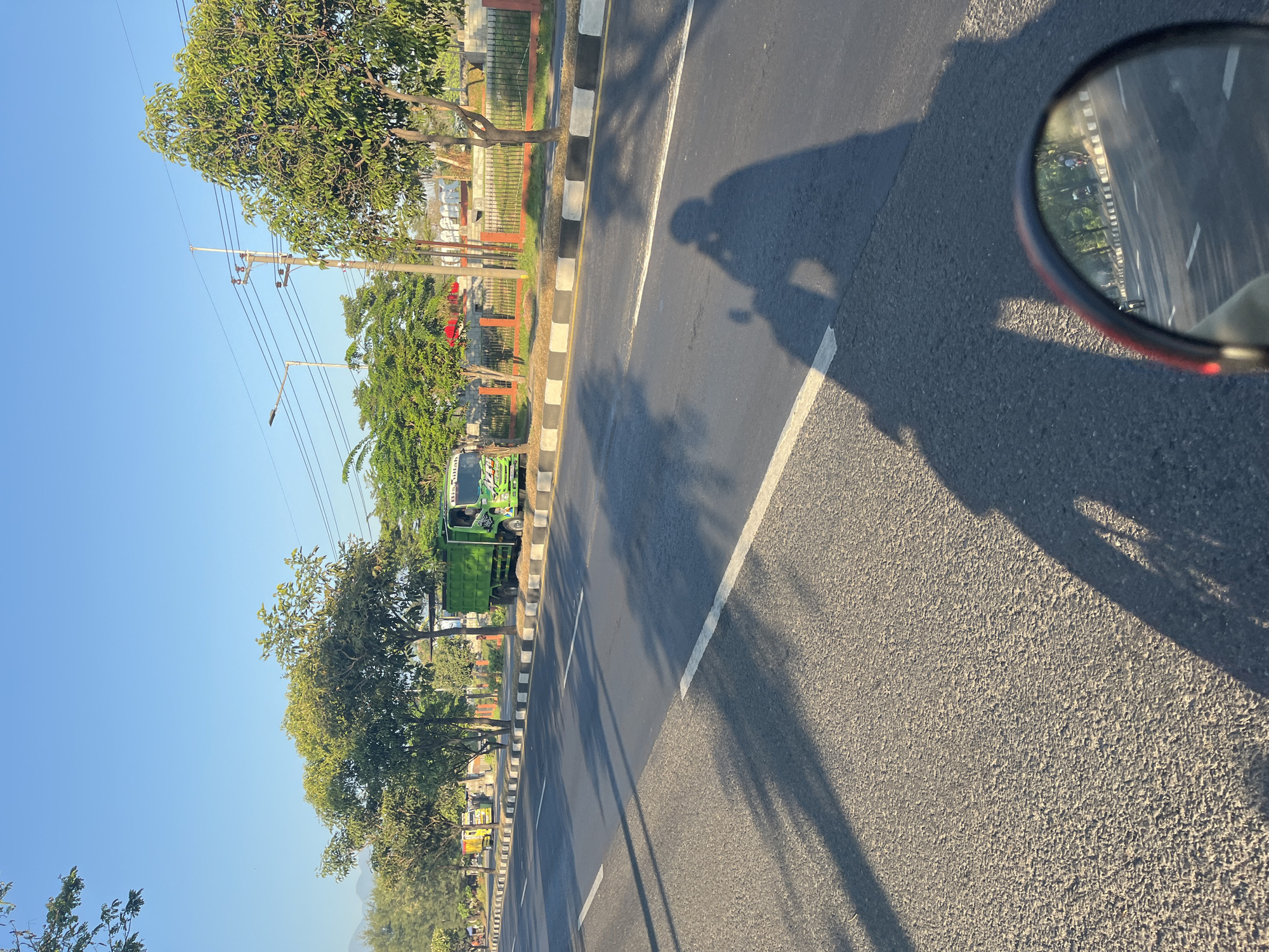 The image depicts a street scene in Siring, Indonesia. A green dump truck is parked on the right side of the road, with a smaller yellow truck in the distance. The road is asphalt, with white and black striped curbs. Trees line the side of the road, casting shadows across the asphalt. In the foreground, the shadow of a person on a motorcycle is visible, along with a motorcycle's rearview mirror reflecting the road ahead. The sky is clear and blue.