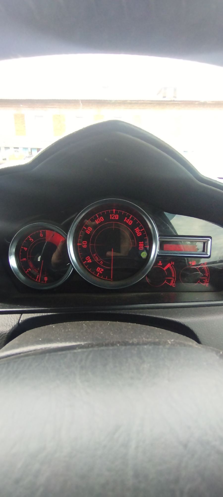 The image shows the interior of a car, focusing on the dashboard with its illuminated red gauges. The speedometer, which indicates speed in km/h, shows a reading of approximately 50 km/h. To the left of the speedometer is a tachometer, indicating engine RPM. To the right of the speedometer, there is a digital display showing "P" for park, along with two smaller circular gauges, one likely for fuel level and the other for engine temperature. The steering wheel is partially visible in the foreground. The dashboard and gauges are illuminated, suggesting it is daytime with the interior lights on or the gauges are inherently backlit. The car appears to be stationary. The background shows a bright, hazy sky and a building with multiple windows and a light-colored facade, indicating an outdoor environment. There is no indication of the location being in Покосное, Russia.