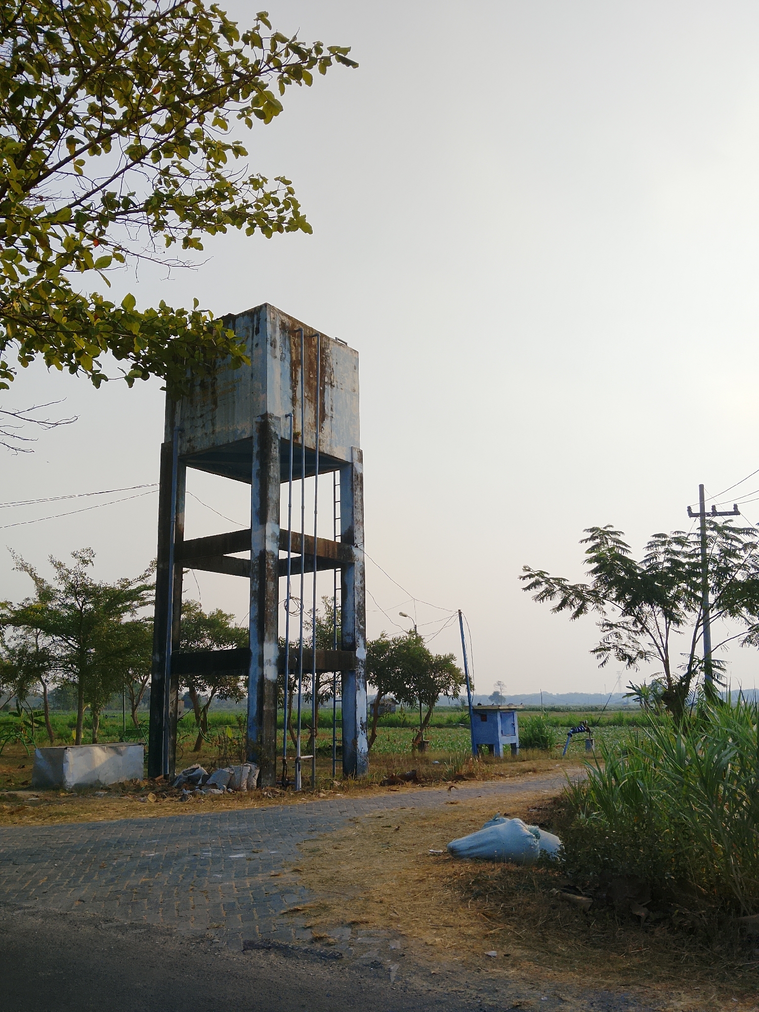 The image depicts an outdoor scene near Ngadrin, Indonesia, dominated by an old, weather-beaten water tower. The tower is tall and square, made of concrete pillars supporting a metal tank that shows significant signs of rust and peeling paint. It stands on a dirt and paved surface, with debris scattered around its base. To the left, tree branches with green leaves hang over the frame. In the background, there are more trees, fields, and a small blue structure with a pump. Electrical poles and wires are visible, indicating the presence of infrastructure. A light blue tarp-covered object lies on the ground in the foreground, near tall green grass. The sky is bright and hazy, suggesting it is daytime. There are no people visible in the image.