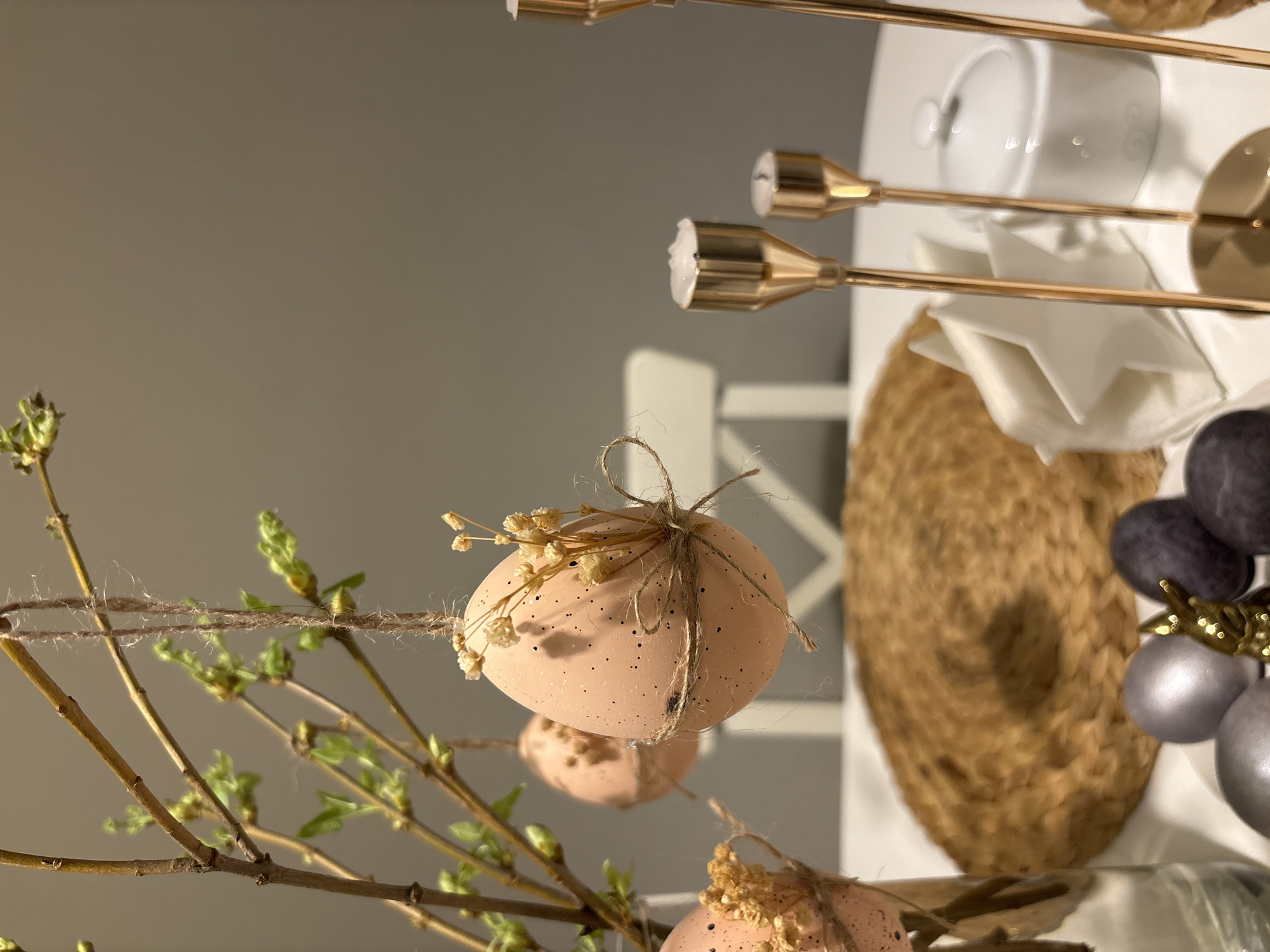 The image shows an Easter decoration consisting of painted eggs suspended from a twig. The eggs are peach-colored with brown speckles, tied with twine, and decorated with dried flowers. The twig has small green buds. In the background, there is a blurred arrangement of objects on a white surface, including a gold candelabra, a white bowl, a woven placemat, and decorative spheres. The scene appears to be indoors, possibly in a domestic setting. The location, Chaikovsky, Russia, is not discernible from the visual information in the image.