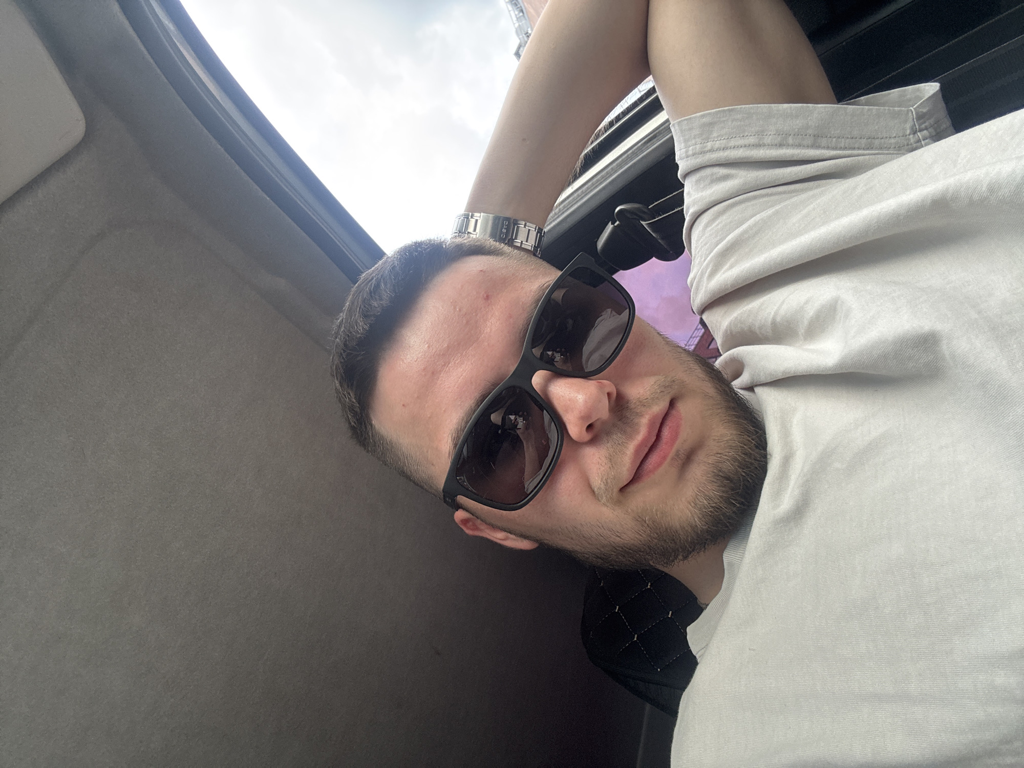 A man in his early twenties, with short dark hair and a light beard, is seen from the chest up, wearing sunglasses and a light-colored t-shirt. He is inside a vehicle, as indicated by the car's interior visible in the background, including the headliner and a portion of the dashboard. The man's left arm is resting behind his head, and he is looking at the camera. The background shows a slightly overcast sky, and the overall lighting suggests it is daytime. The image appears to be a selfie taken within a car.
