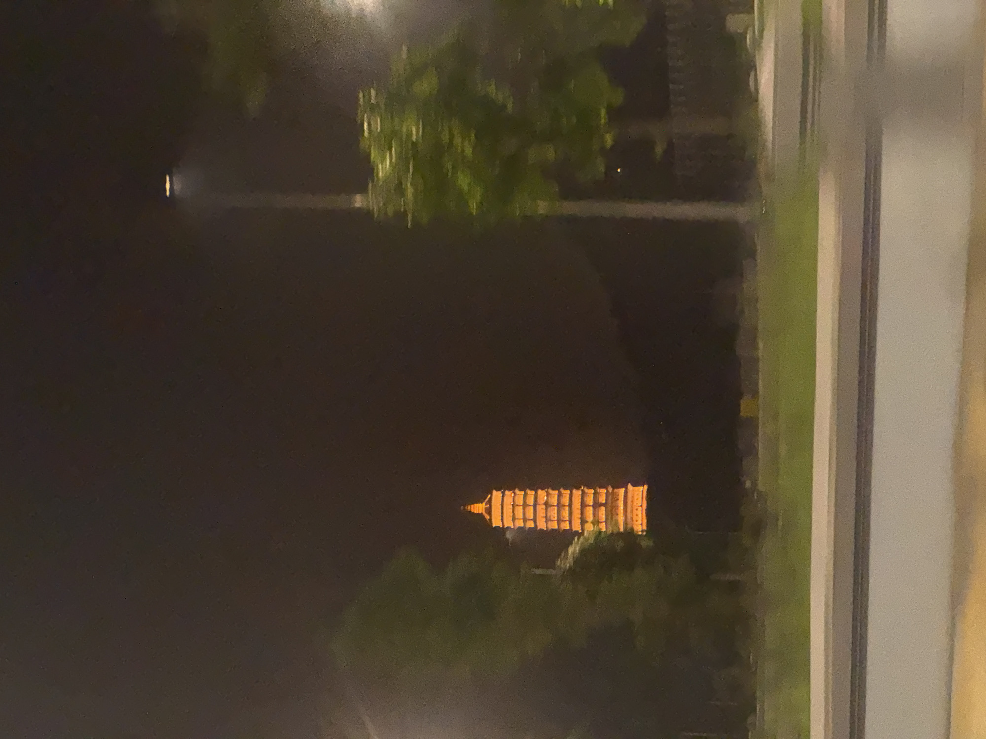 A night time photograph captures a pagoda, illuminated by orange lights, in the distance. The pagoda has a stepped, multi-tiered structure, with a pointed roof. Surrounding the pagoda are dark trees and foliage. In the foreground, a street light with an illuminated head stands to the right of the pagoda, also casting a warm glow. A road with a grey barrier runs horizontally across the bottom of the image, with a strip of green grass and more foliage visible behind it. The sky is dark, suggesting it is late evening or night. The overall scene is one of an urban landscape at night, with a prominent architectural landmark. This pagoda is likely the Trấn Quốc Pagoda, a historical and cultural site located in the Tây Hồ District of Hanoi, Vietnam, known for its beauty and spiritual significance.