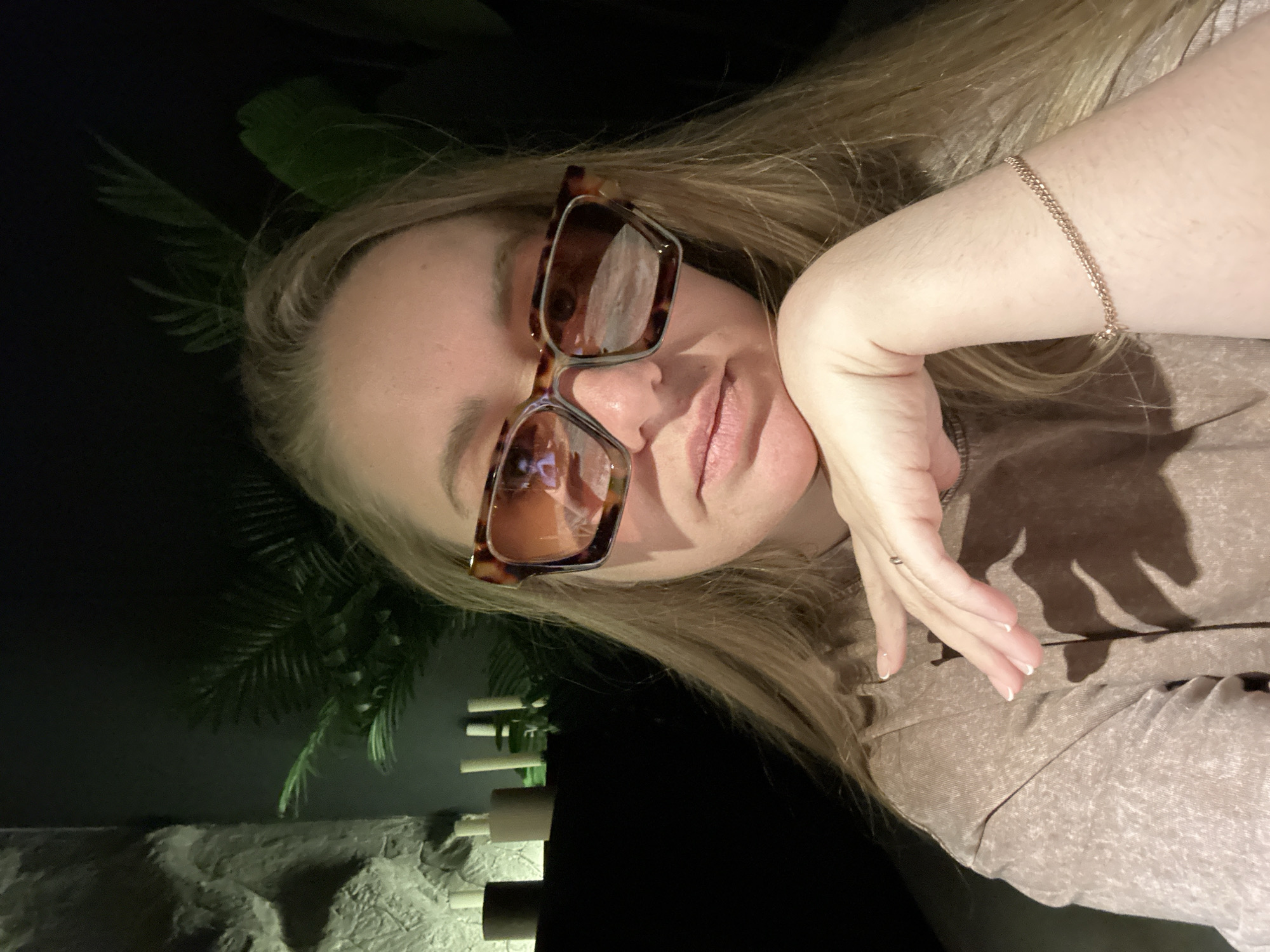 The image depicts a fair-skinned female subject taking a selfie in a dimly lit indoor environment within the city of Chaikovsky, Russia. The subject has long, straight, blonde hair and is wearing large, tortoiseshell-patterned sunglasses with amber-tinted lenses, which reflect a bright light source. She is dressed in a light brown, textured short-sleeved top. Her right hand is positioned with fingers curled, supporting her chin and jawline; a simple gold-colored ring is visible on her right ring finger. A thin, double-chain, rose-gold-colored bracelet is worn on her right wrist. The background features a textured, light-colored wall or panel illuminated by a cool, green-tinged light on the left. Below this, a dark surface displays several decorative cylindrical objects, including two dark and several light-colored elements, possibly candles or holders. Green foliage is visible behind these objects and extends into the dark right side of the background, suggesting potted plants. The overall lighting is dim, with the subject's face well-lit from the front.