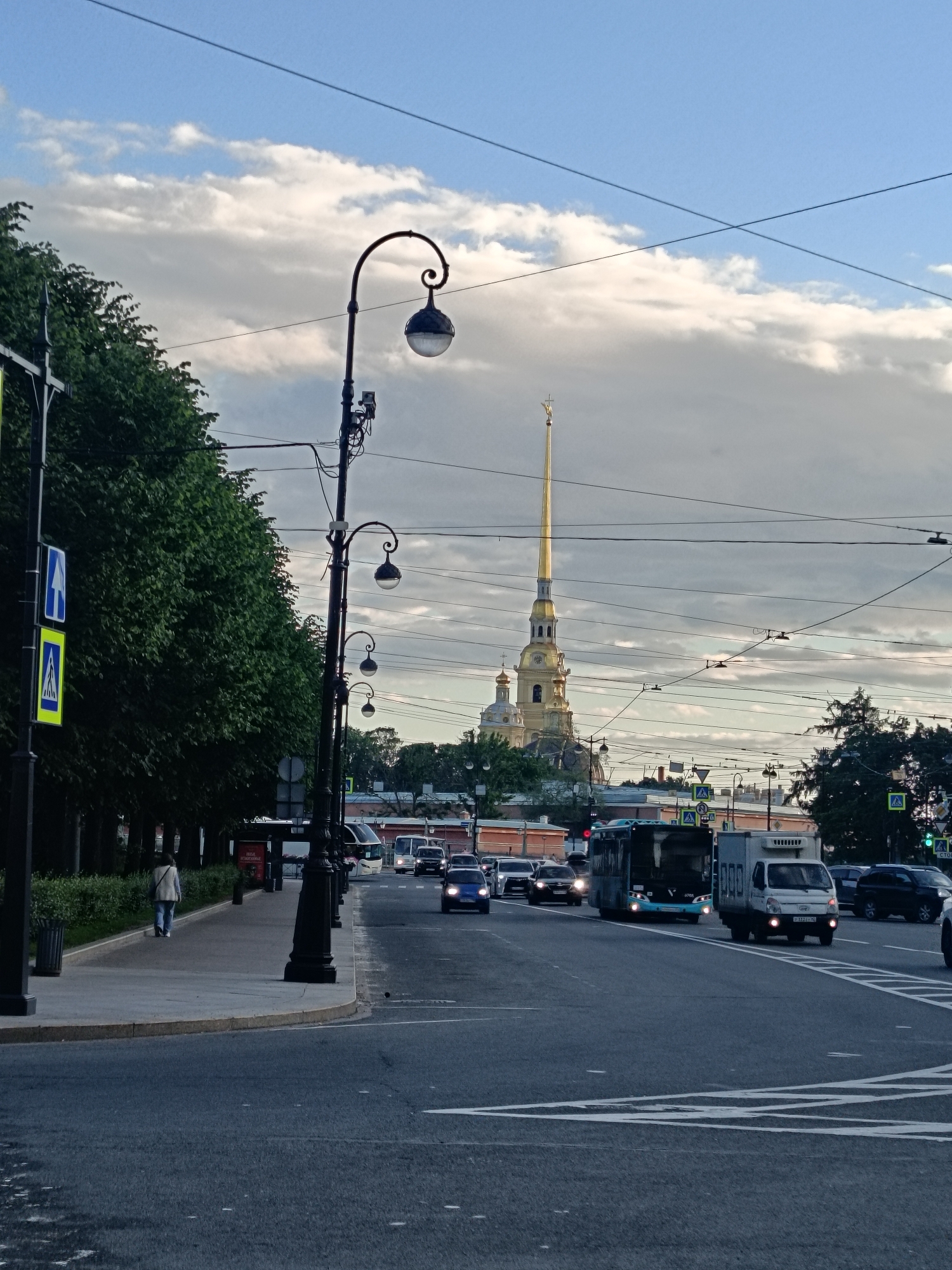 The image depicts a street scene in Saint Petersburg, Russia, featuring a prominent spire of a golden-topped building in the background. The sky is partly cloudy, suggesting it is either early morning or late afternoon. The foreground shows a paved road with several vehicles, including cars and a bus, in motion. A pedestrian walkway runs alongside the road, bordered by lush green trees. A lone figure is seen walking on the sidewalk. Several ornate streetlights line the walkway, adding to the urban ambiance. The overall mood is calm and serene, characteristic of a city in the transitional hours of the day. No visible text is discernible in the image.
